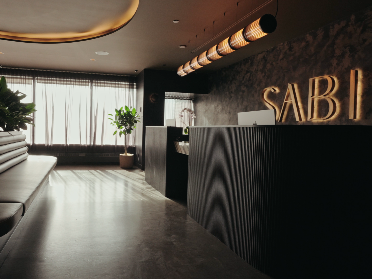 Modern medical clinic with black textured walls, neon glowing sign spelling 'SABI', elegant reception desk, leather bench seating, potted plants, large windows with sheer curtains, and warm ambient lighting.