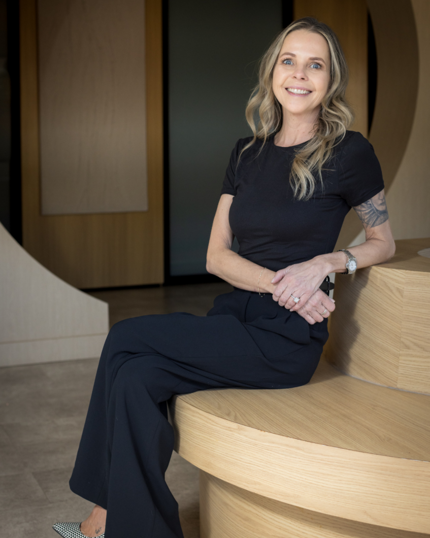 A woman with blonde wavy hair, wearing a black short-sleeved shirt and black pants, sitting on a curved wooden bench in an indoor space, smiling at the camera.