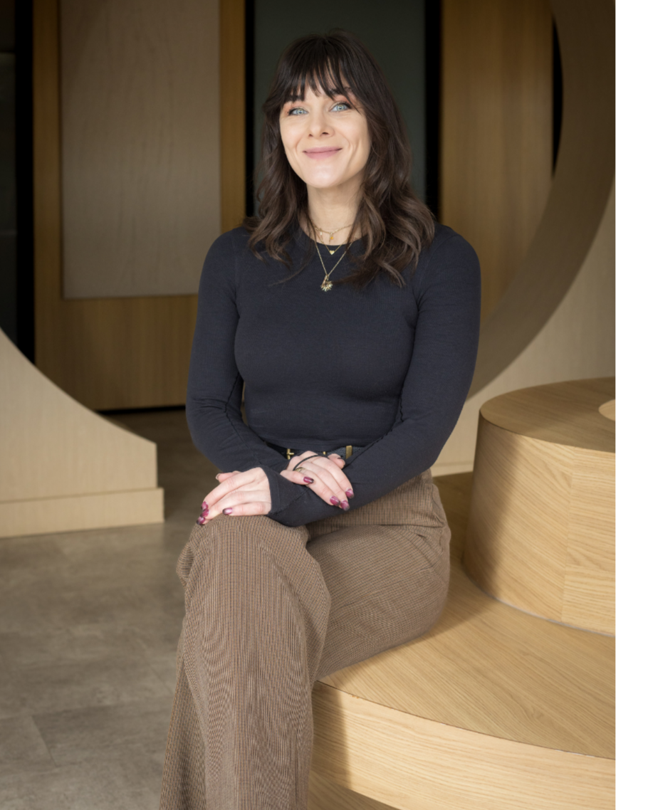 A woman with dark brown hair and bangs smiling while sitting on a curved wooden bench in a modern indoor setting.