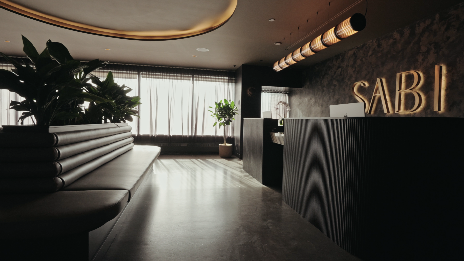 Modern medical spa reception area with black partition, potted plants, window curtains, and illuminated "SABI" sign on the wall.