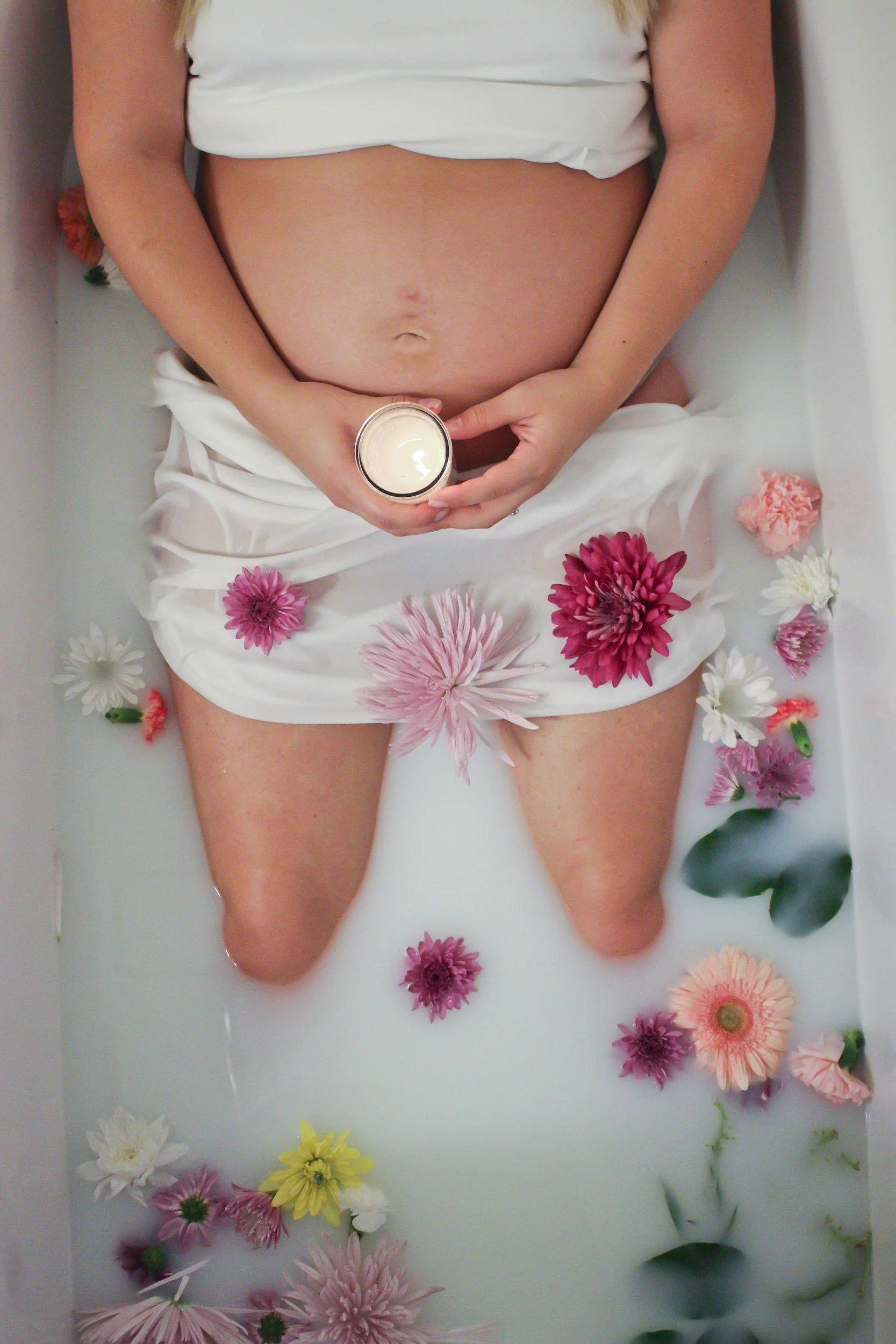 A person lying in a milk bath surrounded by colorful flowers, with hands holding a lit candle.