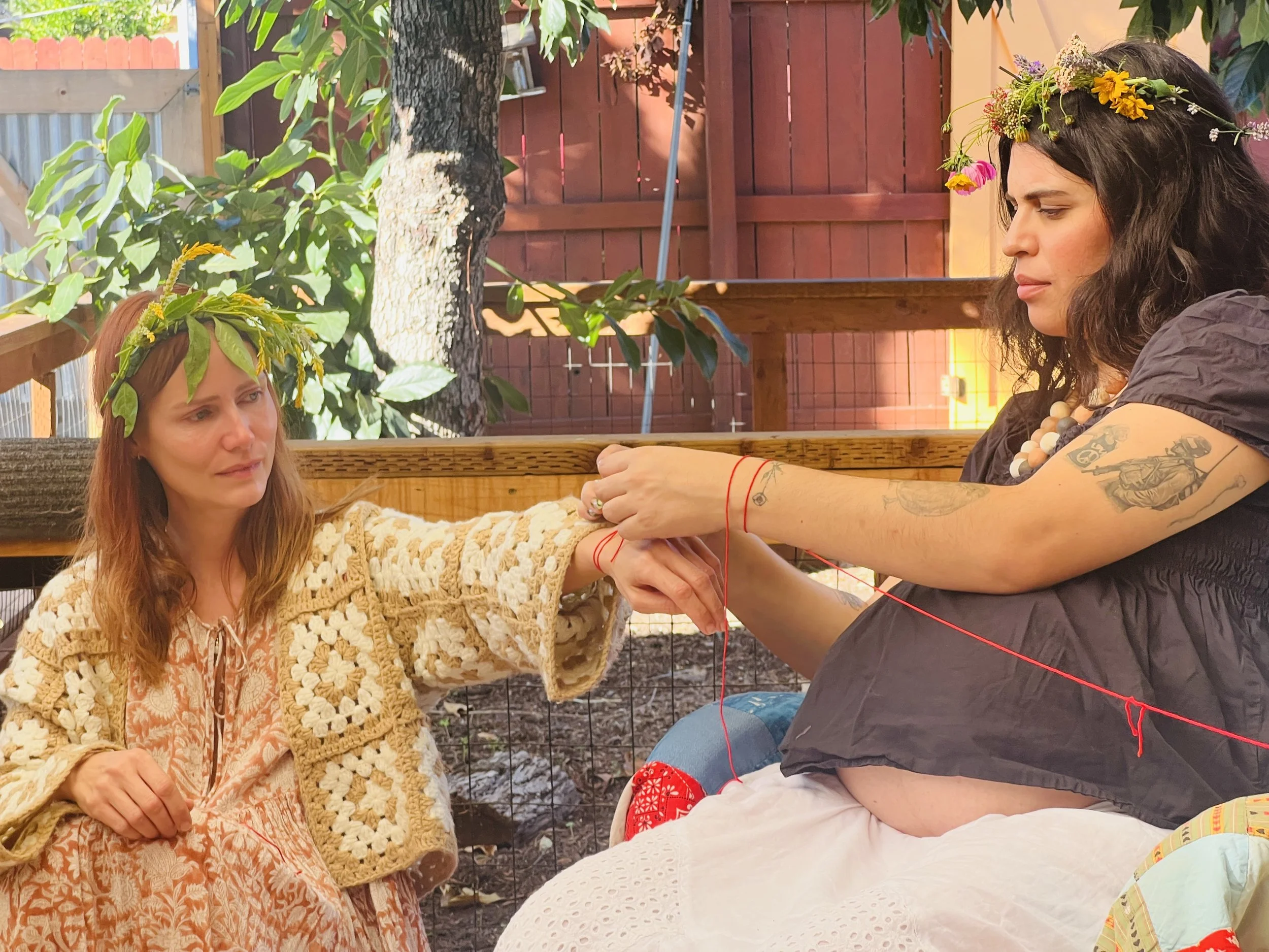 Two women are sitting outdoors, engaged in a yarn-begging craft. One woman with long red hair and a floral crown made of leaves and yellow flowers looks at the other woman. The other woman, with dark hair and a crown of mixed flowers, is focused on the craft, which involves red yarn. The background shows a wooden fence, some green foliage, and a tree.