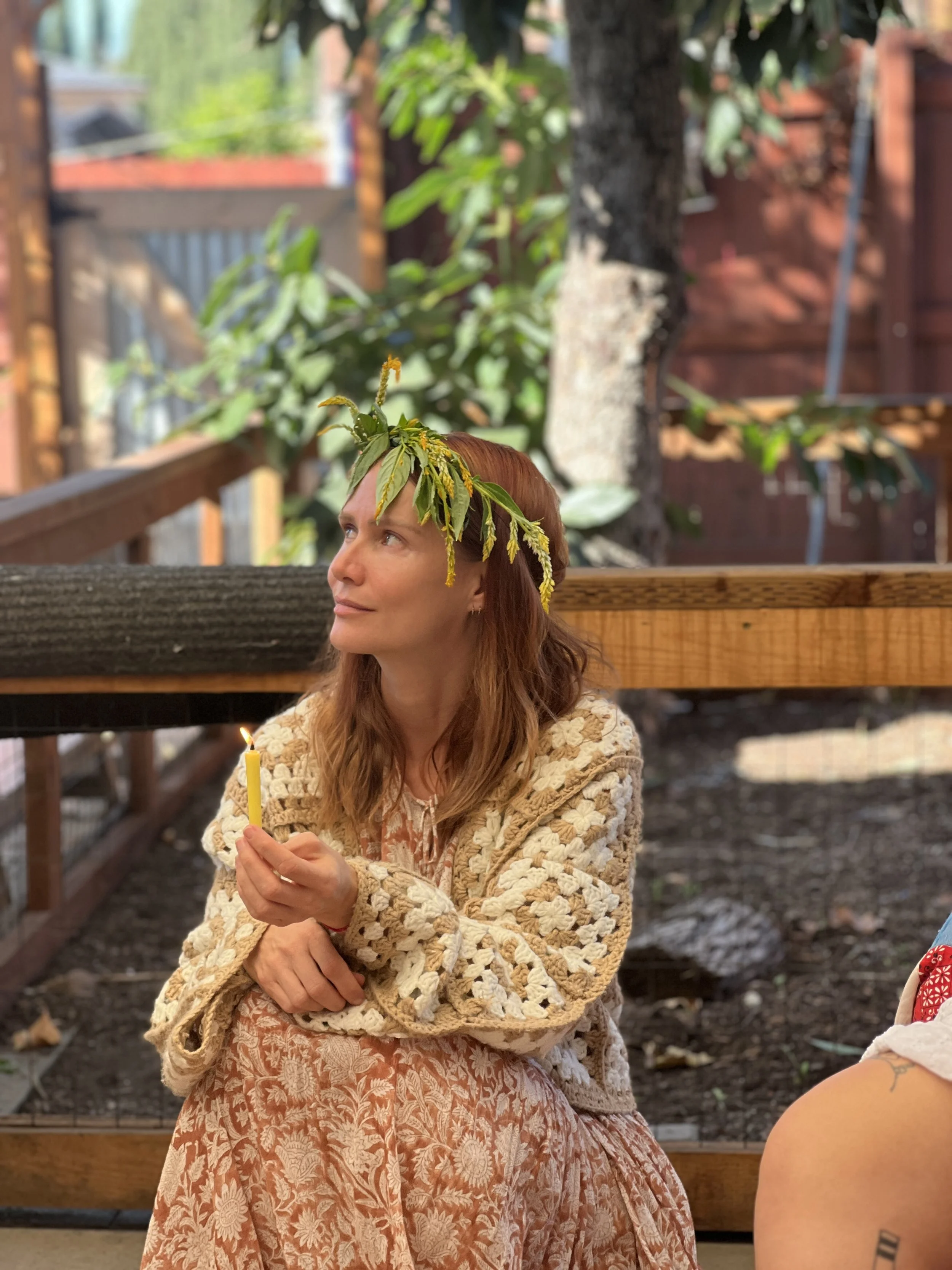 A woman with reddish hair, wearing a floral dress and a crocheted cardigan, holding a lit candle. She has a wreath of greenery and yellow flowers on her head. She is outdoors in a garden or backyard setting, with a wooden fence and trees in the background.