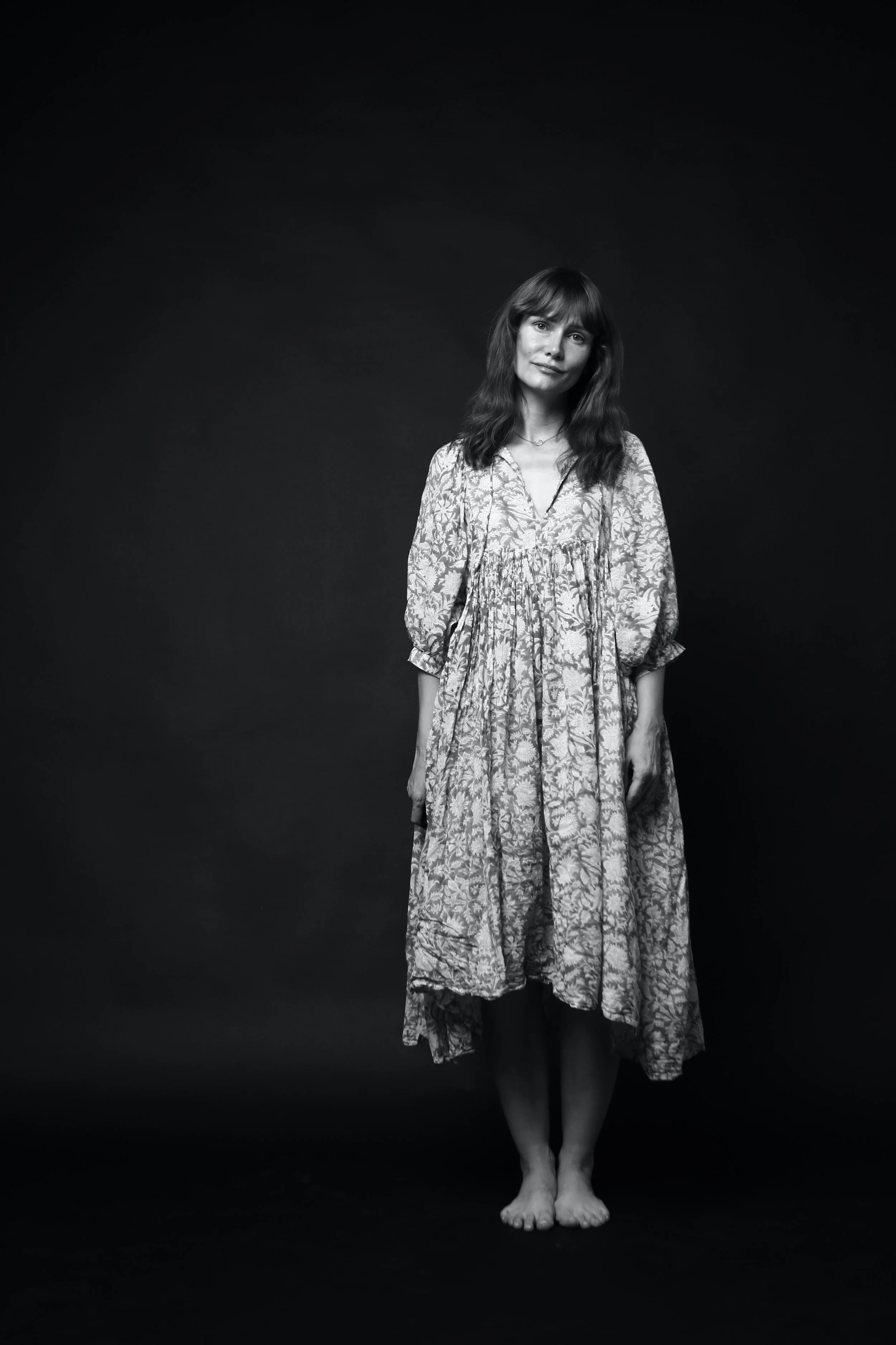 Black and white photograph of a woman standing barefoot against a dark background, wearing a patterned dress, with shoulder-length hair, looking at the camera with a slight smile.
