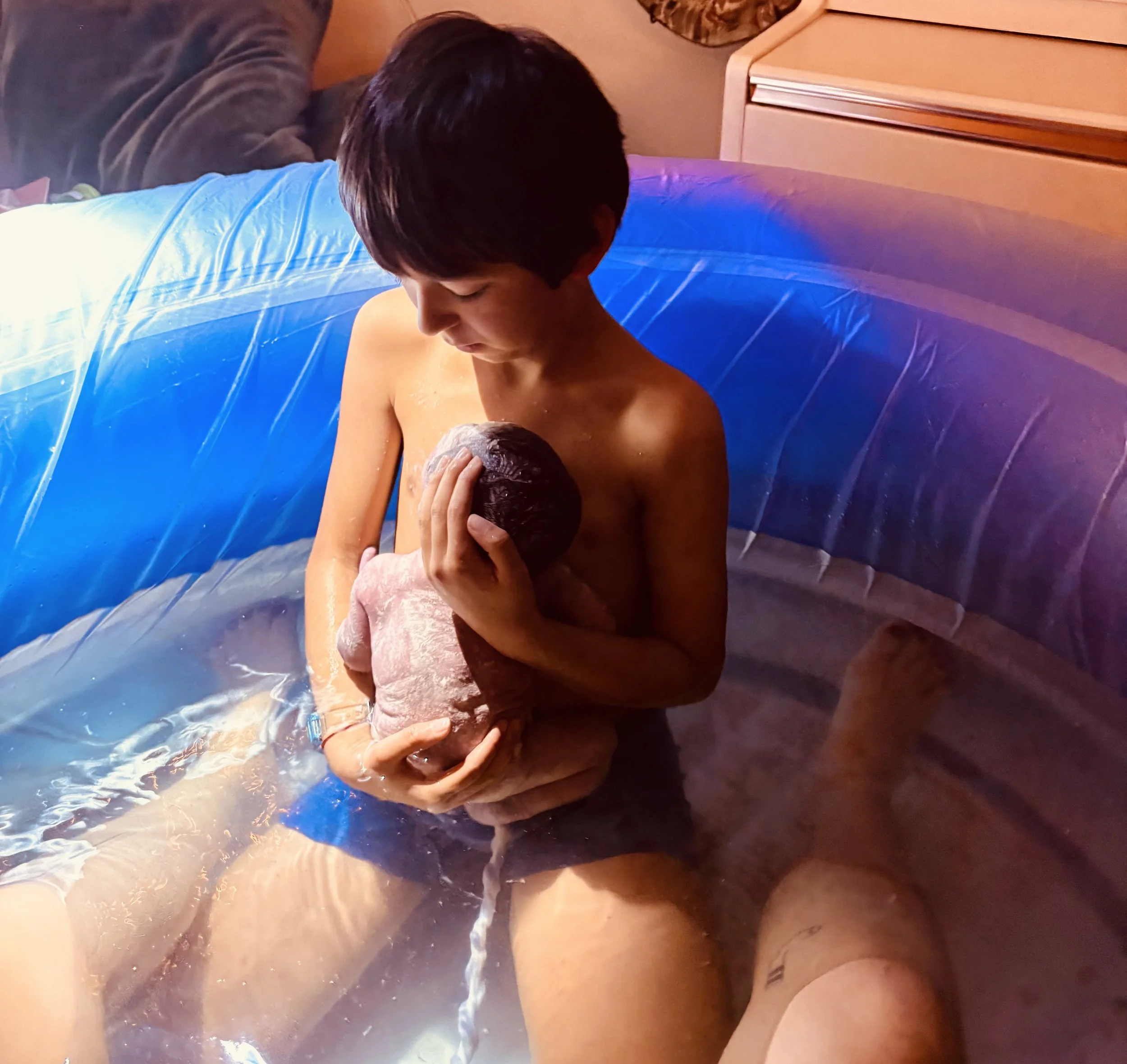 A person with short dark hair swimming underwater with a baby, both looking at each other. The person is holding the baby, and they are in a pool with blue water and a swimming float.