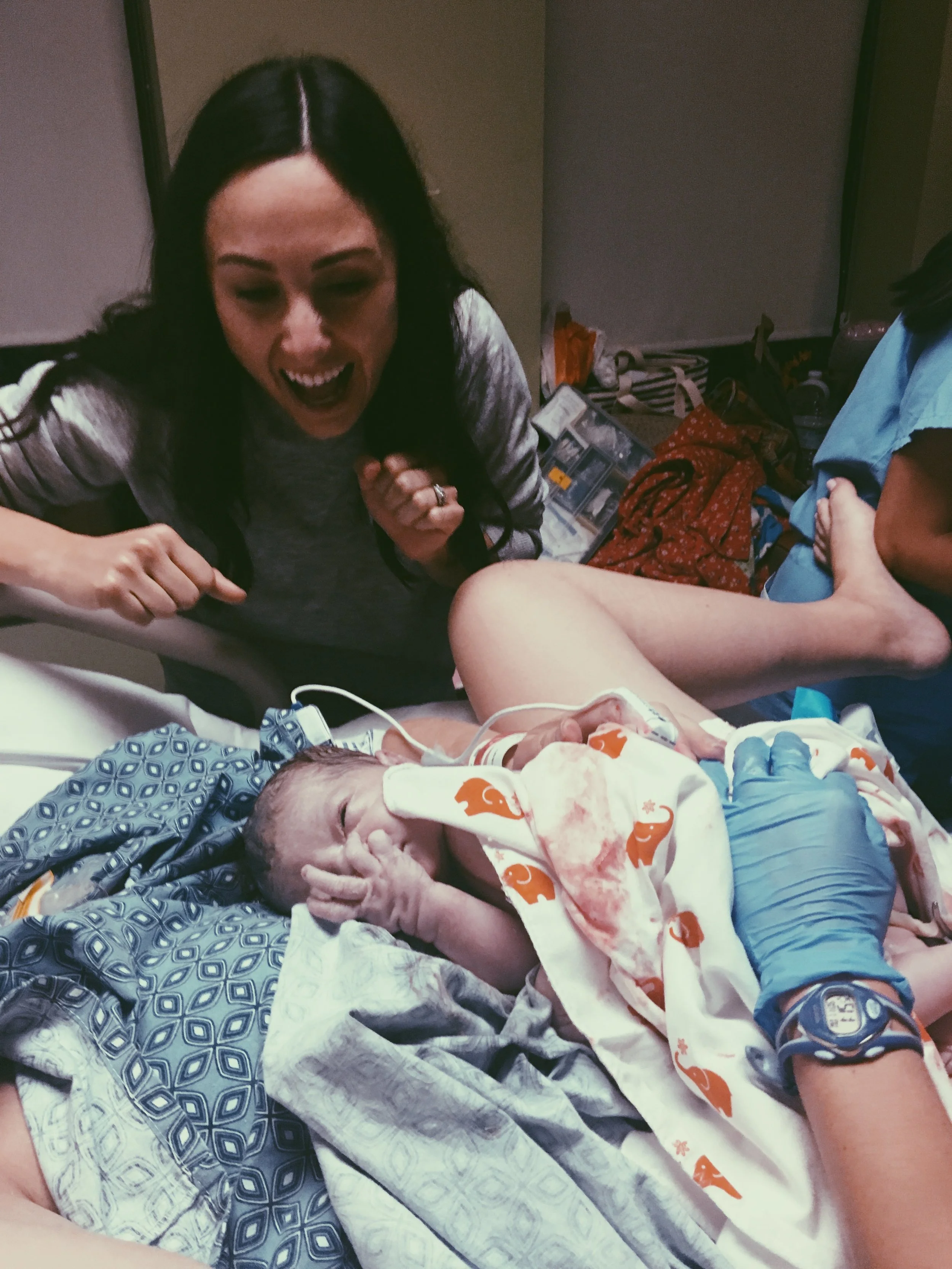 A woman with long dark hair smiling, lying on a hospital bed with a newborn baby. The baby is wrapped in a blanket with orange elephants and has medical tubes attached, in a hospital setting.