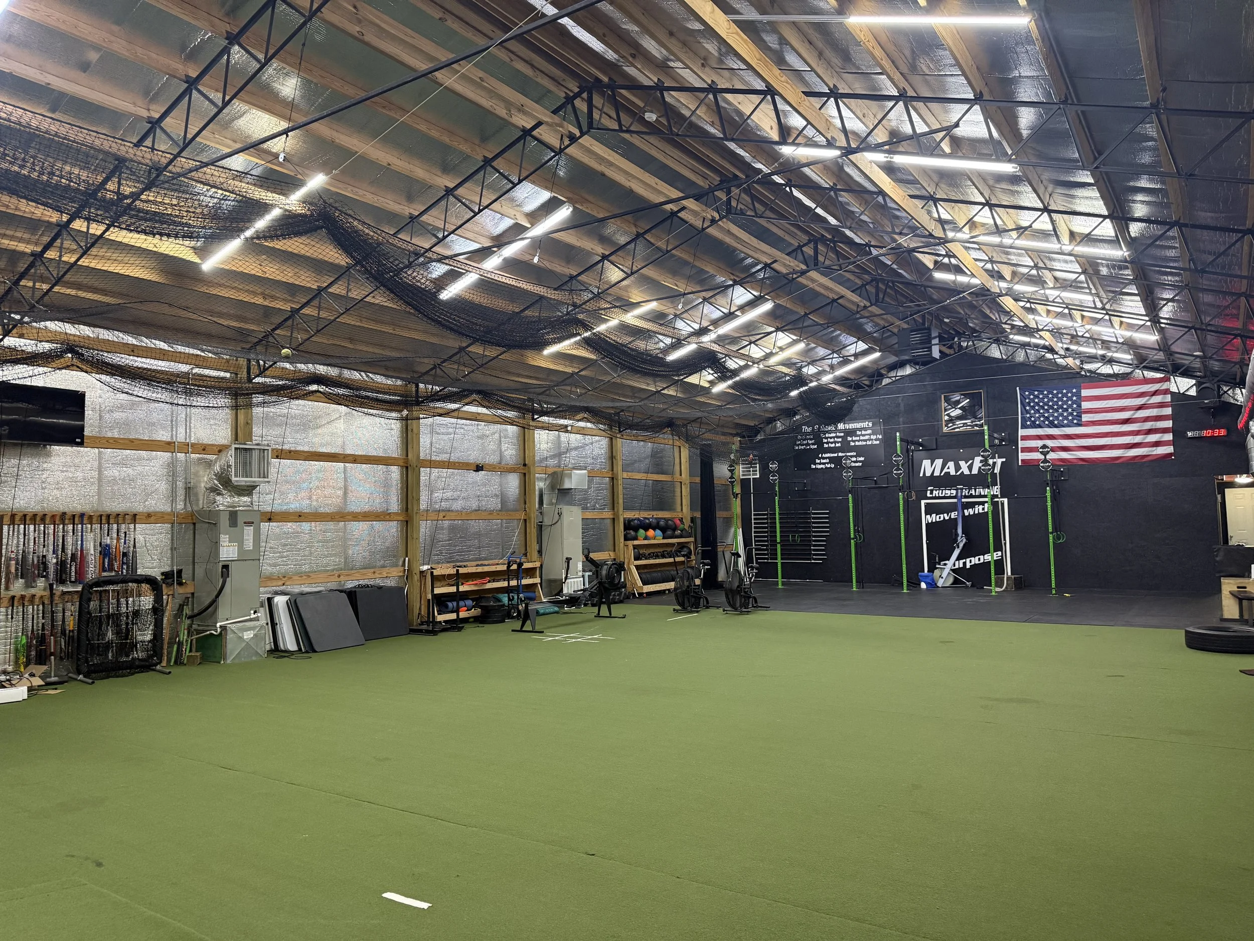 Indoor facility with a green turf floor, black training equipment, and a large American flag on the wall. The ceiling has exposed wooden beams and metal trusses with bright lights.