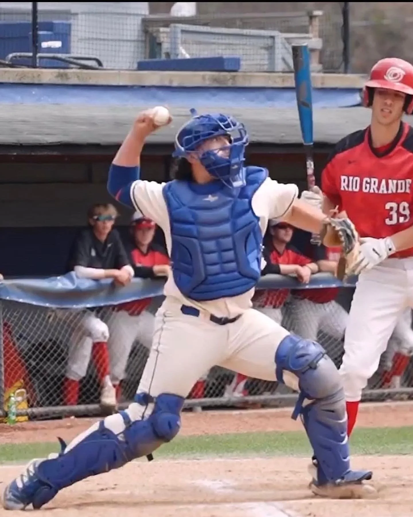 A baseball catcher in blue gear throws a ball during a game at a baseball field, with teammates and officials in the background.