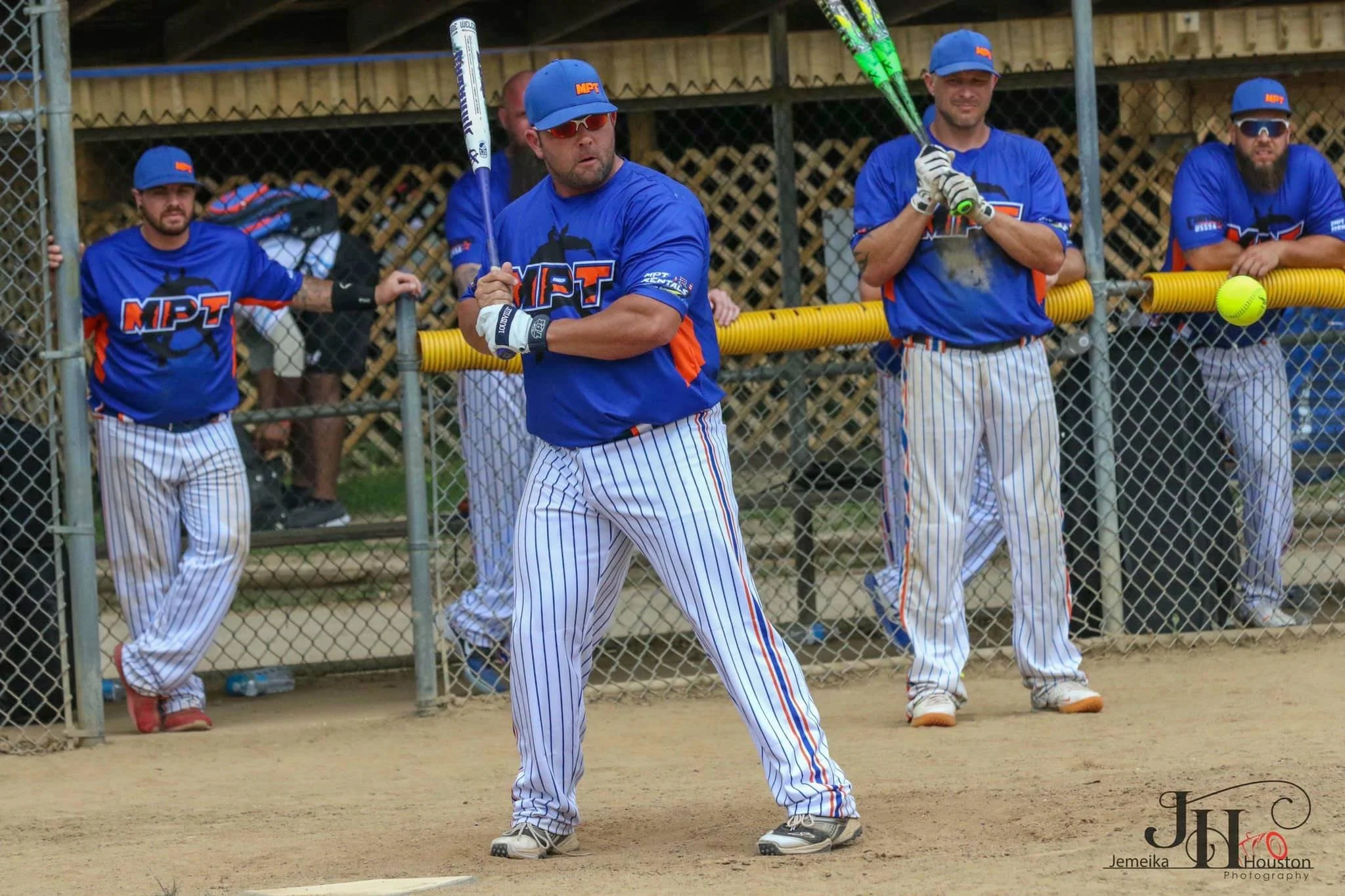 Group of baseball players in blue jerseys and striped pants standing near a fence on the sidelines of a baseball field, with one player holding two green baseball bats and wearing sunglasses, and others watching or waiting.