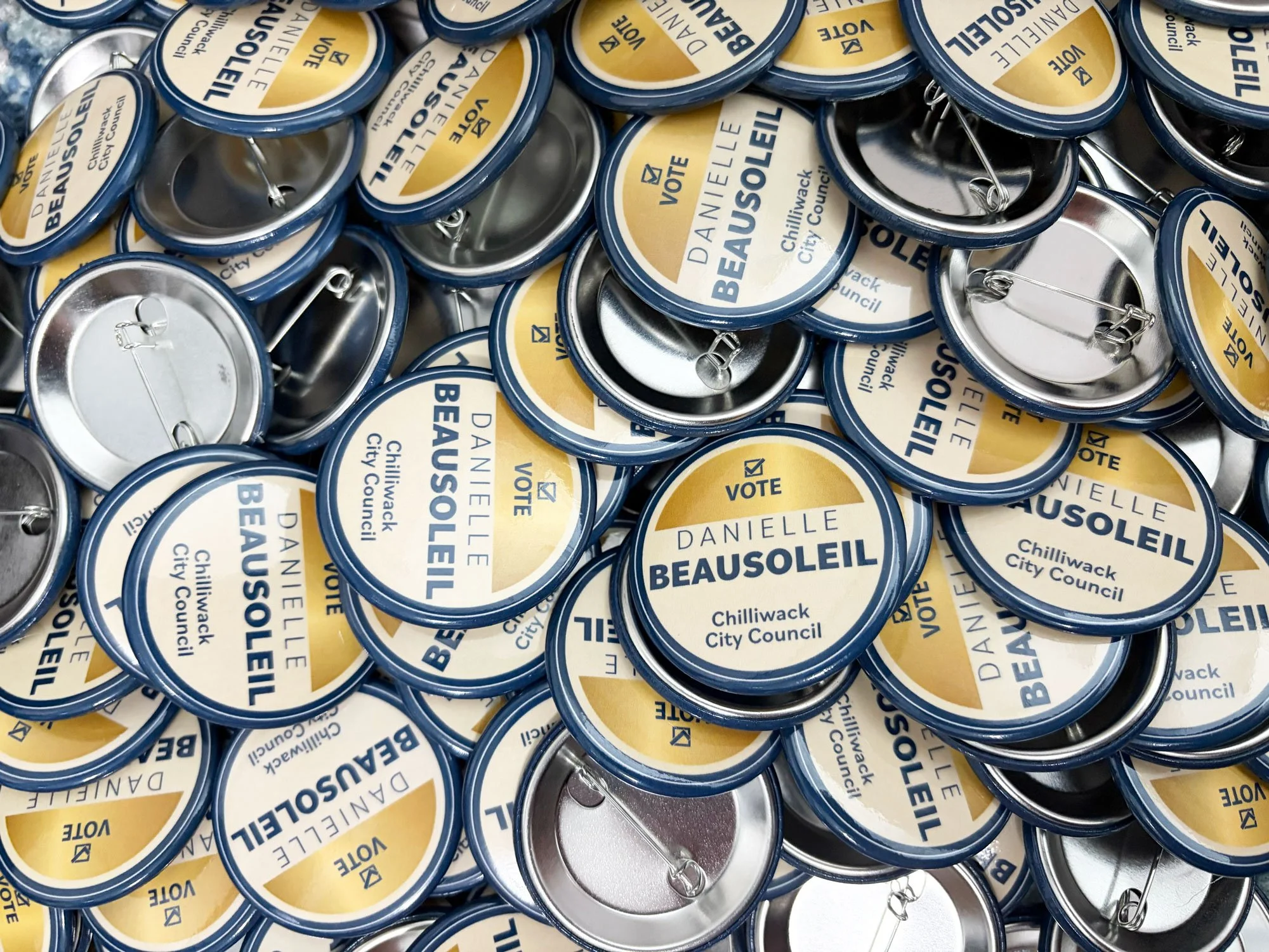 A pile of campaign buttons promoting Danielle Beausoleil for Chilliwack City Council, with a blue and yellow design and the word 'VOTE' on each.
