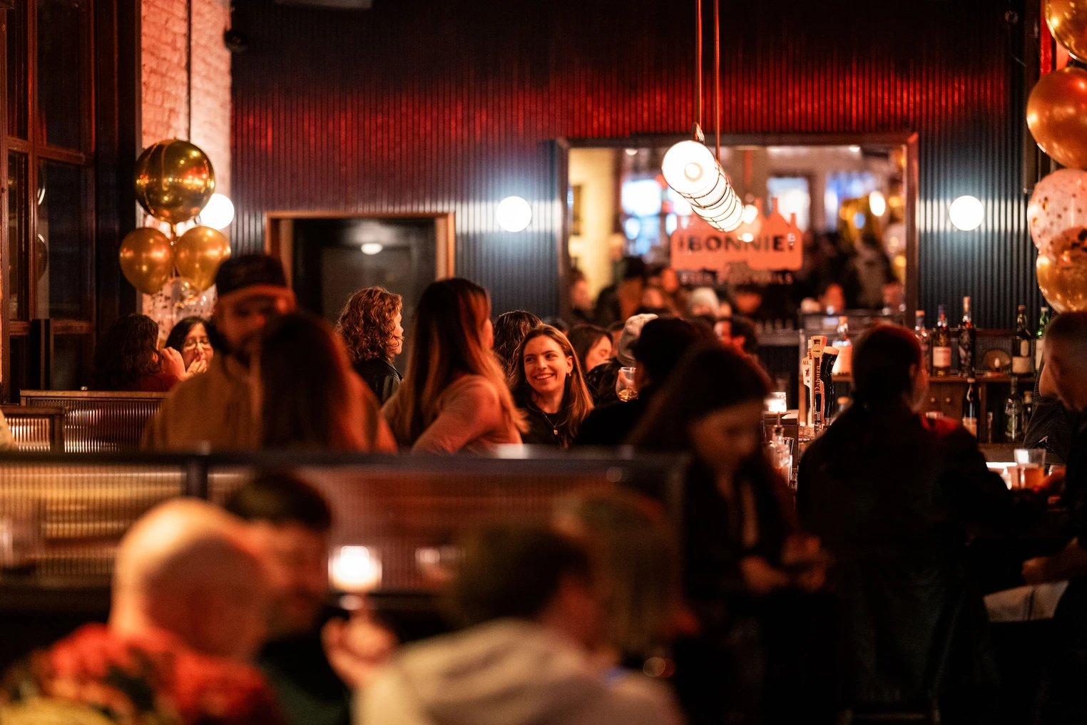 People socializing and enjoying drinks at a lively bar or restaurant with red lighting, balloons, and a bar counter.
