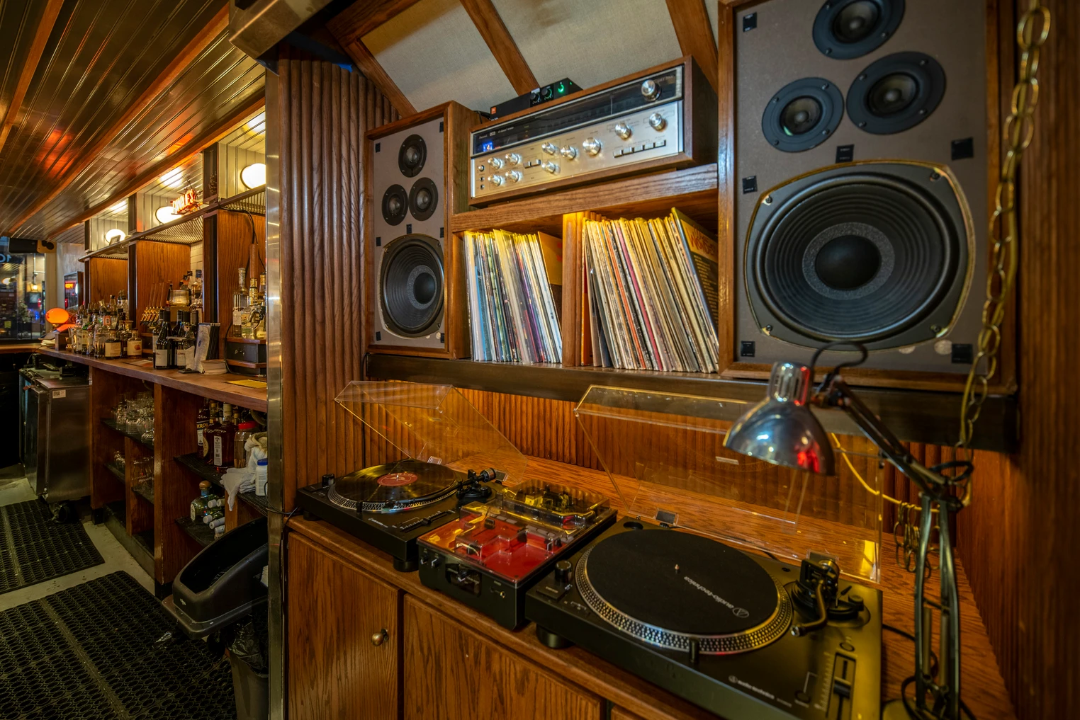 A vintage bar with wooden shelving, a DJ setup with turntables, vinyl records, and large speakers.