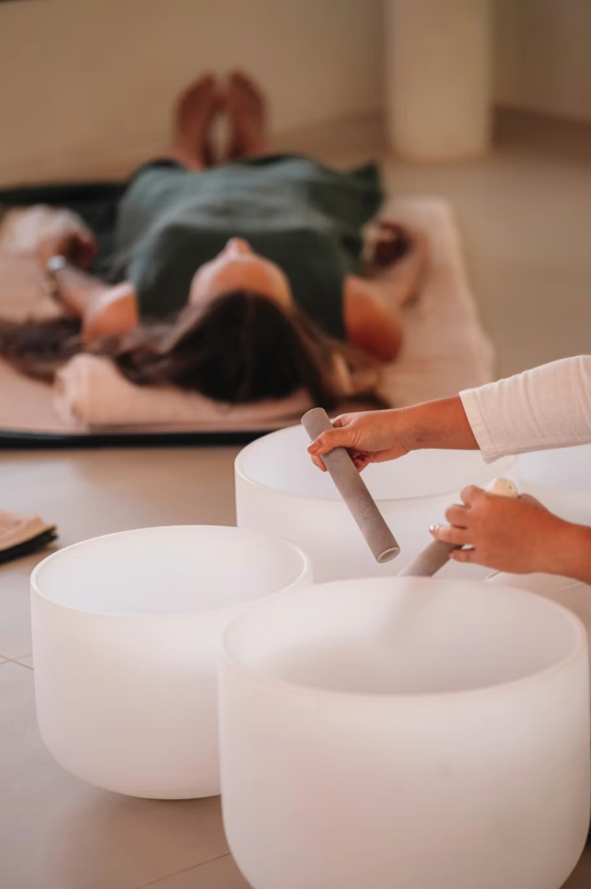 Person lying on a table with arms extended, while another person plays a musical instrument with a mallet in front of large white singing bowls.