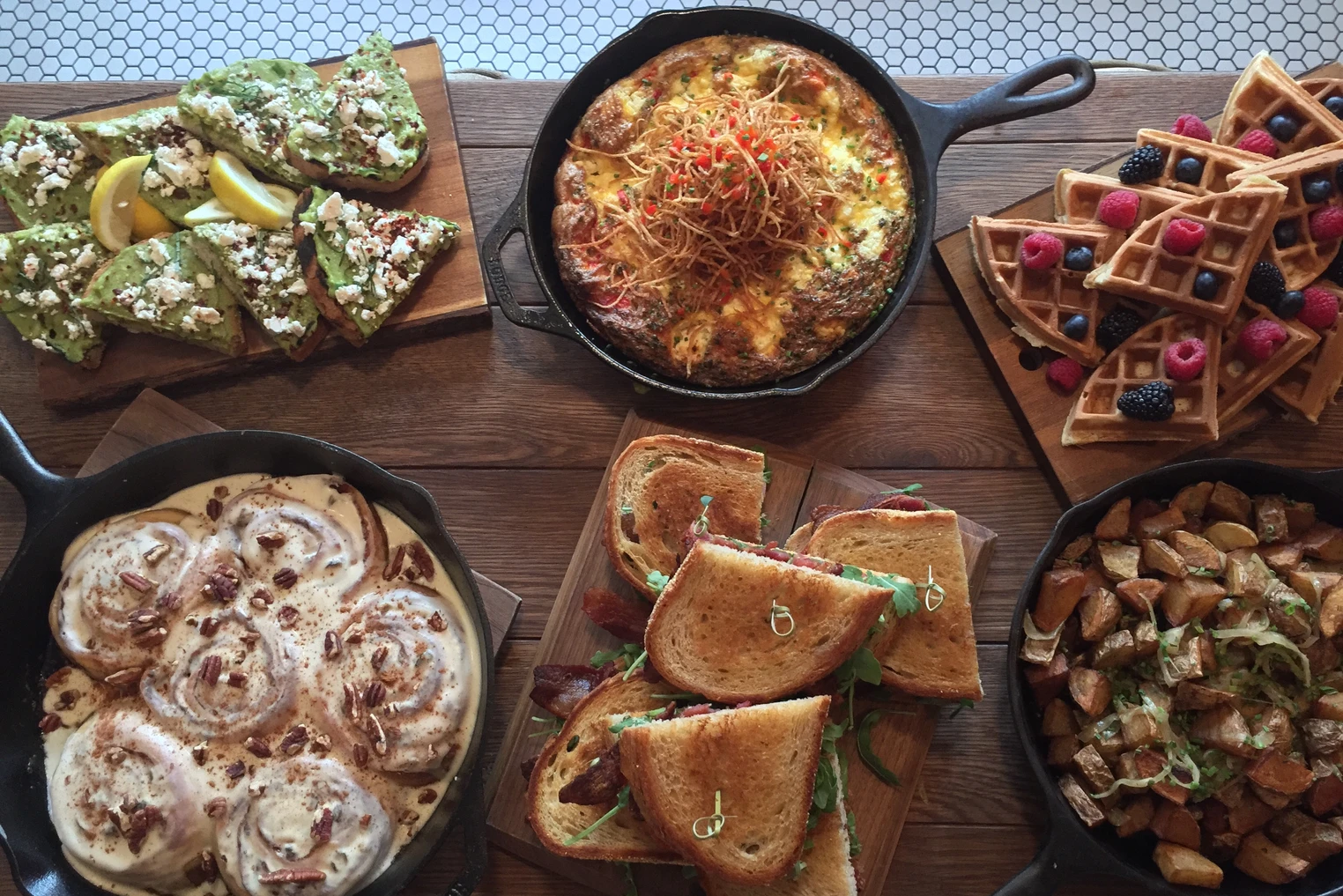 A variety of dishes on a wooden table, including avocado toast, a baked dish with cheese and herbs, waffle sticks with berries, cinnamon rolls with pecans, a sandwich with toasted bread and bacon, and a roasted potato and vegetable medley.