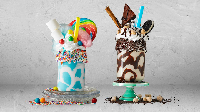 Two colorful milkshakes decorated with candies, cookies, and toppings, placed on stands against a plain background.
