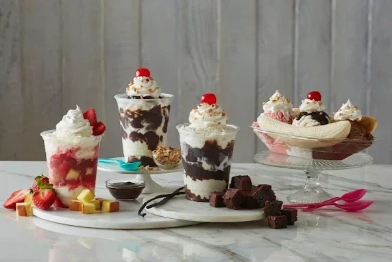 Assorted ice cream sundaes with whipped cream and cherries, some topped with chocolate syrup, served with brownies and strawberries on a white tray and glass platter against a wooden background.