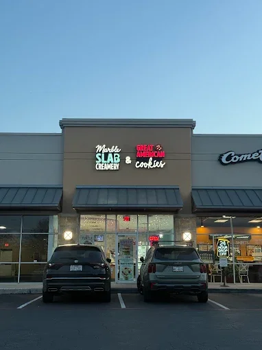 Exterior of a strip mall with a sign for Marble Slab Creamery and Great American Cookies, two parked cars in front, and the store windows showing ice cream and cookie displays.