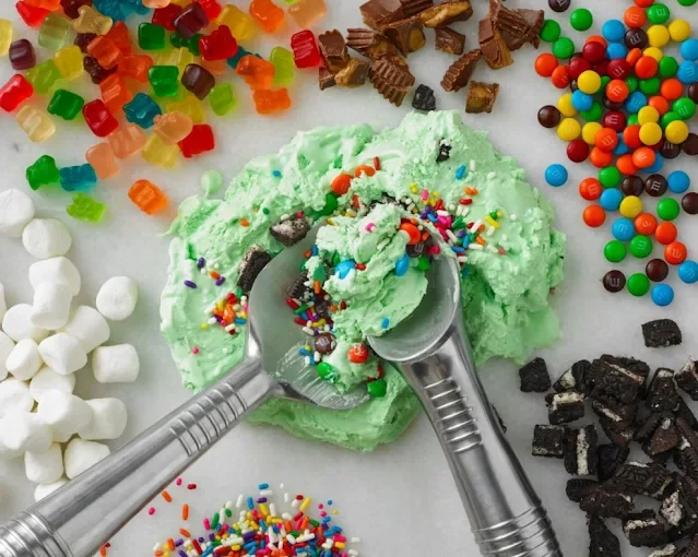 Close-up of a green slime with colorful sprinkles, surrounded by various candies and chocolates on a white surface, with silver scoops.