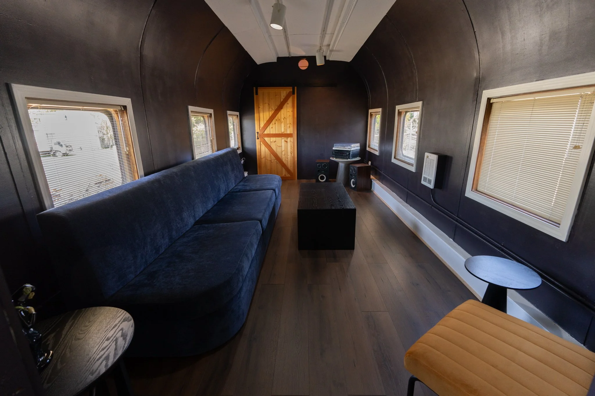 Interior of a small room with dark walls, wooden door, three small windows with blinds, a blue velvet sofa, a black coffee table, audio equipment, speakers, and a small round side table with a yellow cushion.