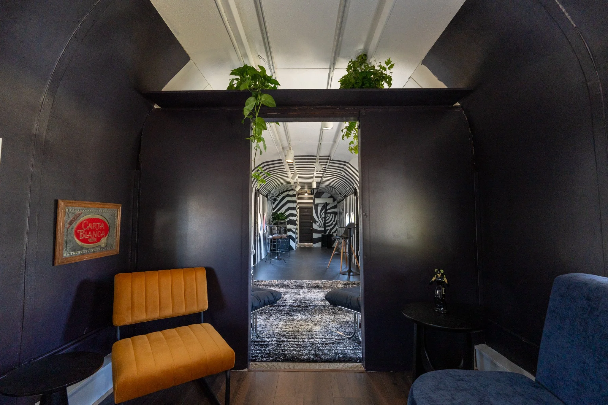 Interior view of a modern, dark-colored lounge with orange chairs, a small round black table, and a framed artwork on the wall. The space extends into a brightly lit corridor with black and white striped walls, plants hanging from the ceiling, and a 