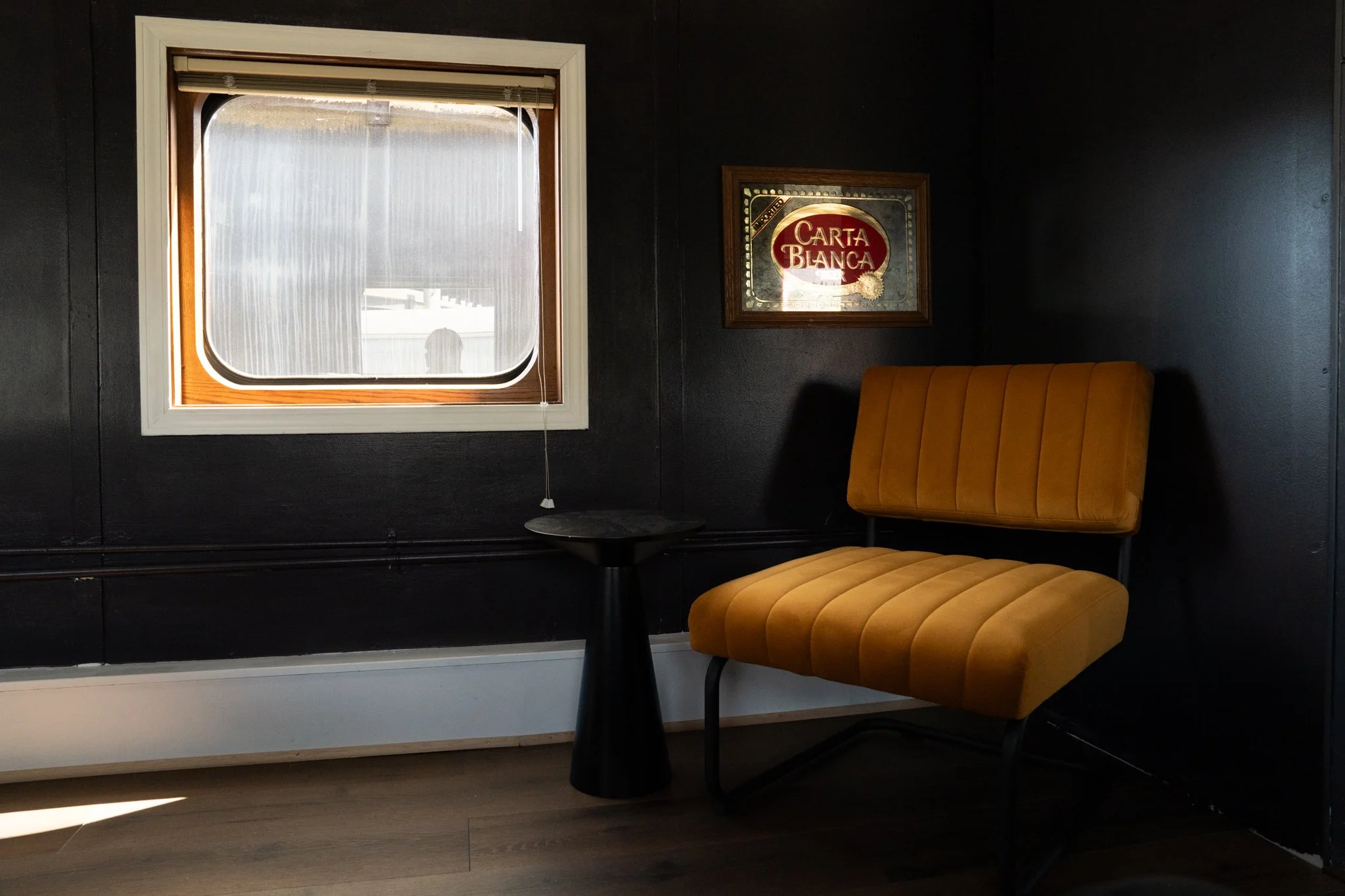 A cozy corner with a window letting in natural light. There is a mustard yellow upholstered chair with vertical tufting and black metal legs, positioned next to a small black round table. The dark wall behind features a framed mirror with the words '