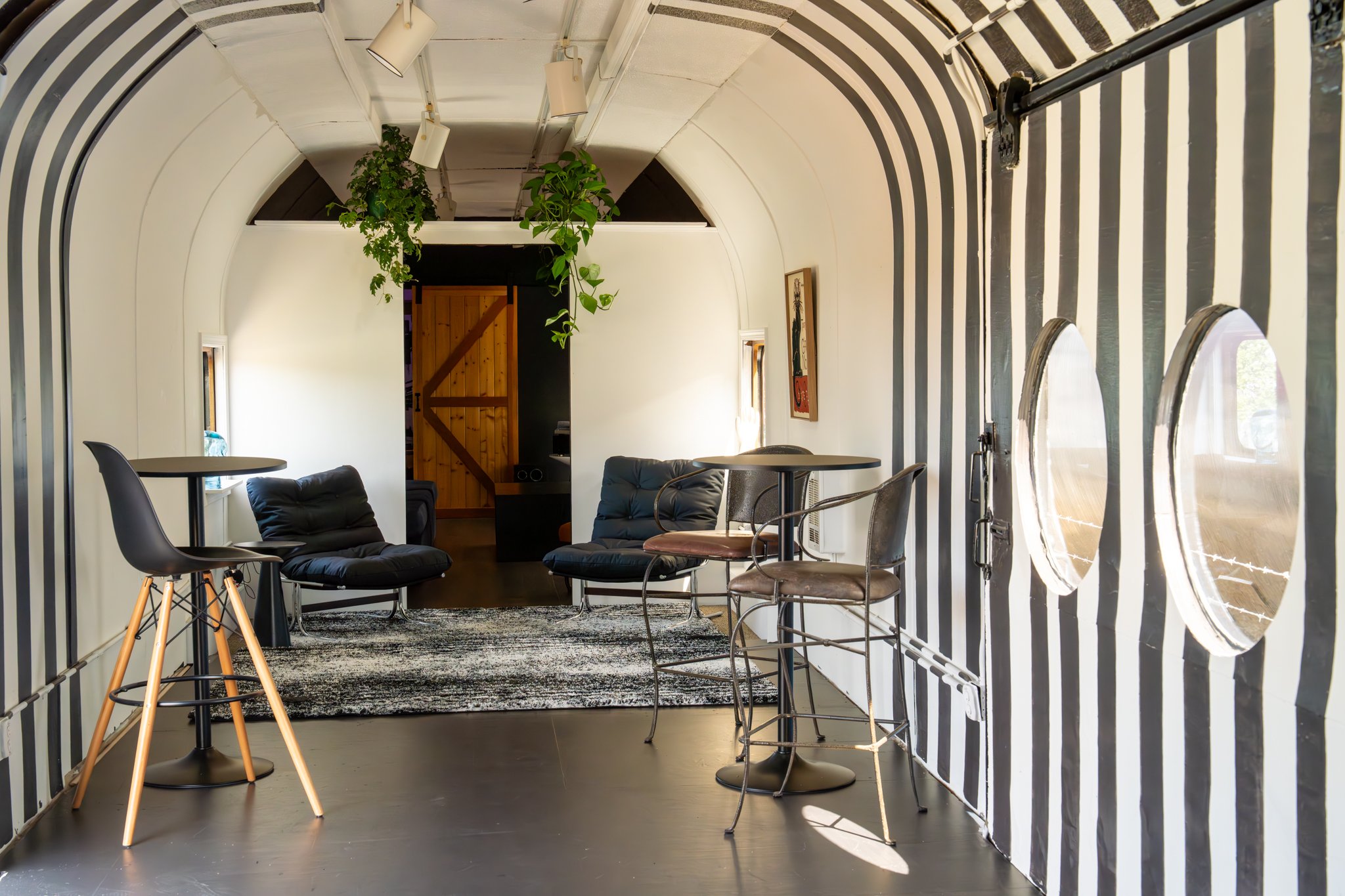 Interior of a uniquely designed room with black and white striped walls, a curved ceiling, and modern furniture including black cushioned chairs, a small round table, and string lights hanging from the ceiling. There are also large circular window op