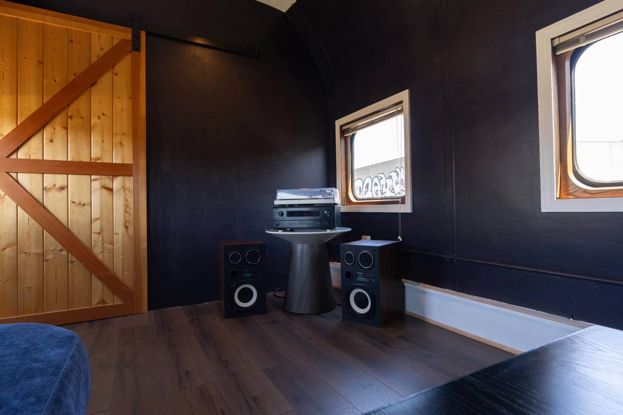 A room with dark walls and two windows, one of which is open, featuring a vintage stereo system and large speakers on a small table.