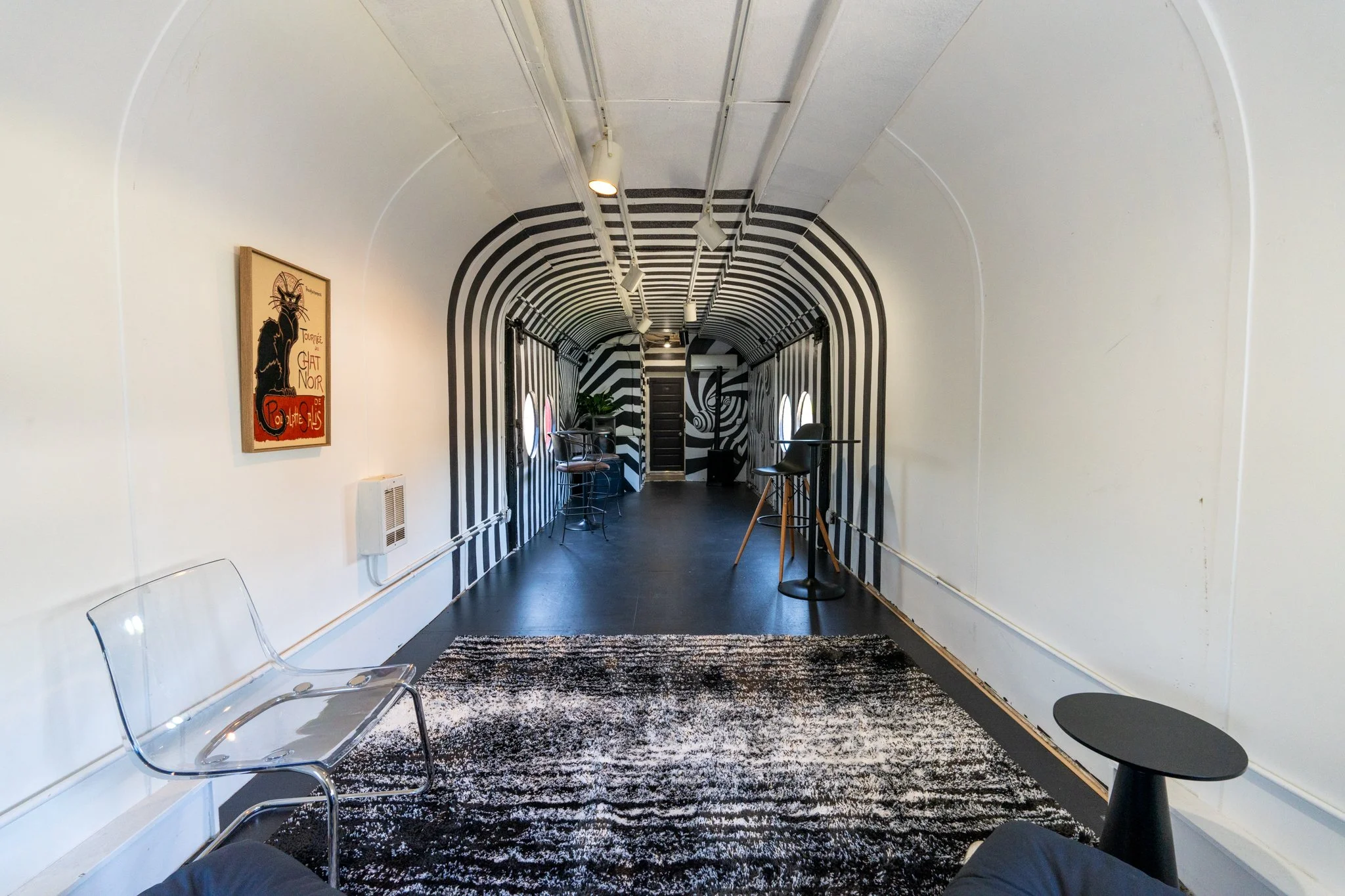 A hallway with black and white striped walls and ceiling, with a curved ceiling design, seen from a traveler's point of view sitting with legs stretched out. There are transparent chairs on the left, a black and white patterned rug, artwork on the wa