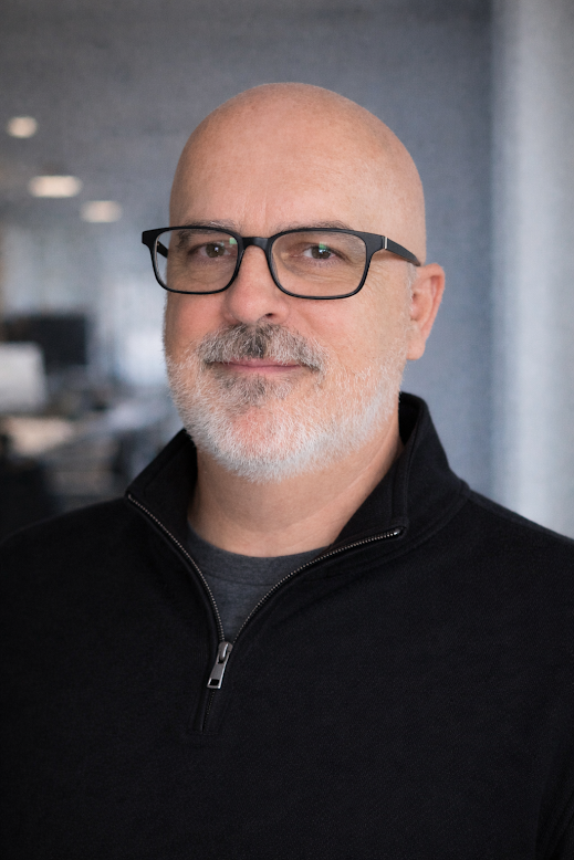A middle-aged man with glasses, a gray beard, and a bald head posing in an indoor setting.