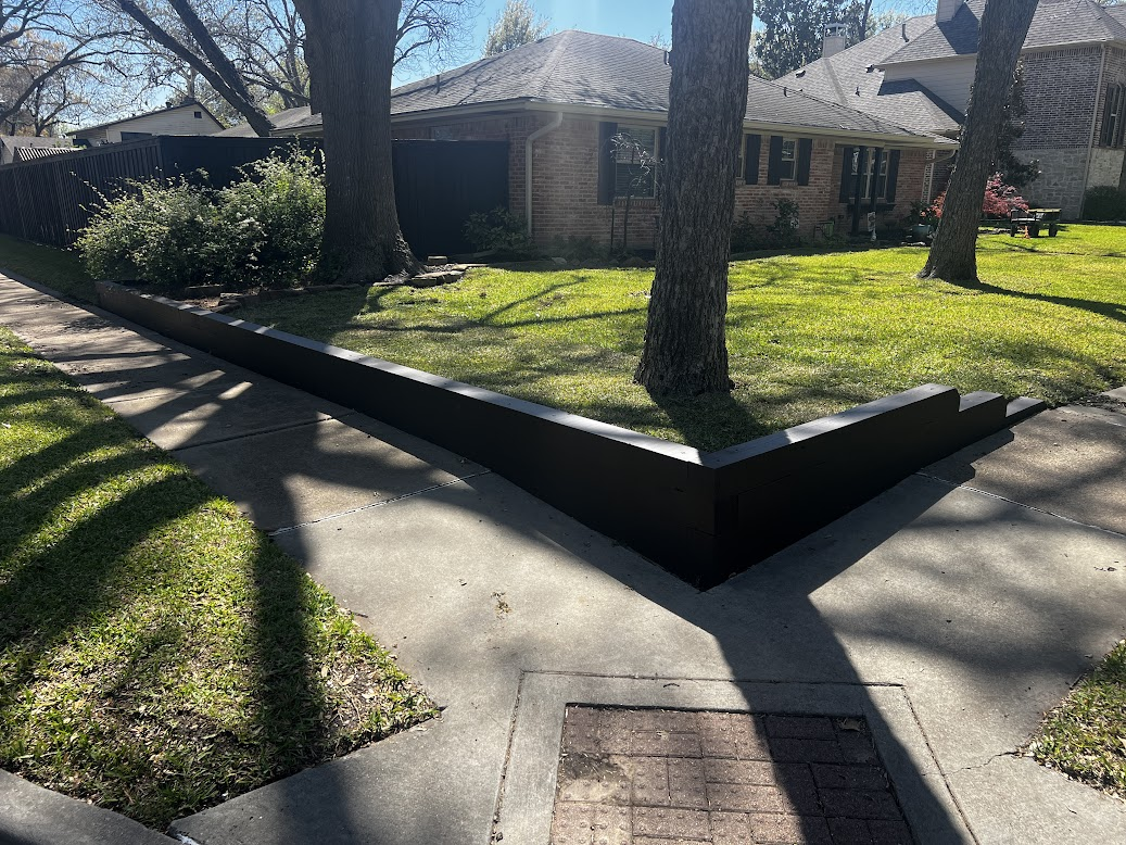 A residential yard with a brick house, trees, a grass lawn, a sidewalk, and a black border wall along the yard edge.
