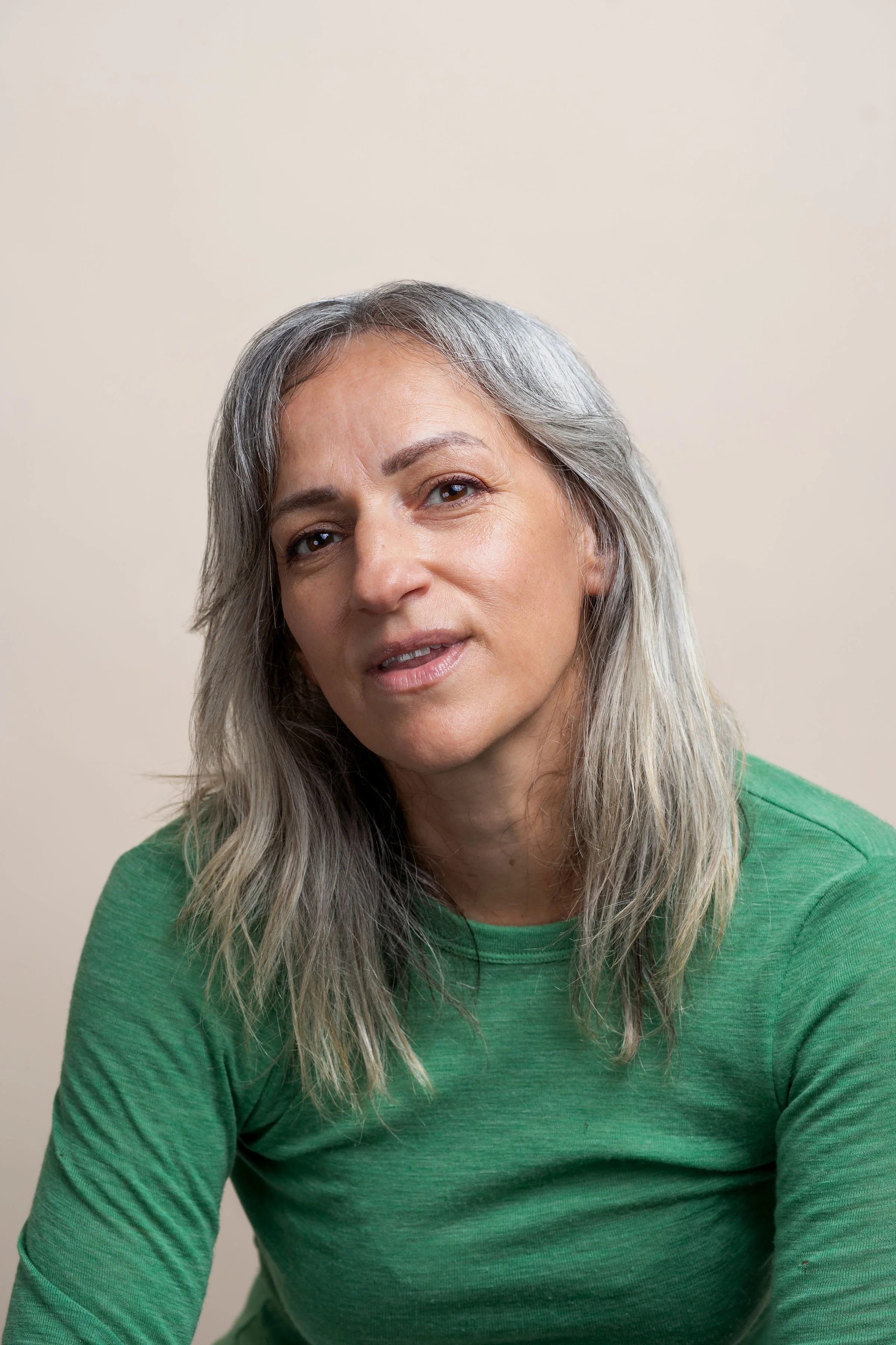 A middle-aged woman with gray hair and a light complexion wearing a green shirt, looking at the camera with a slight smile, against a plain beige background.