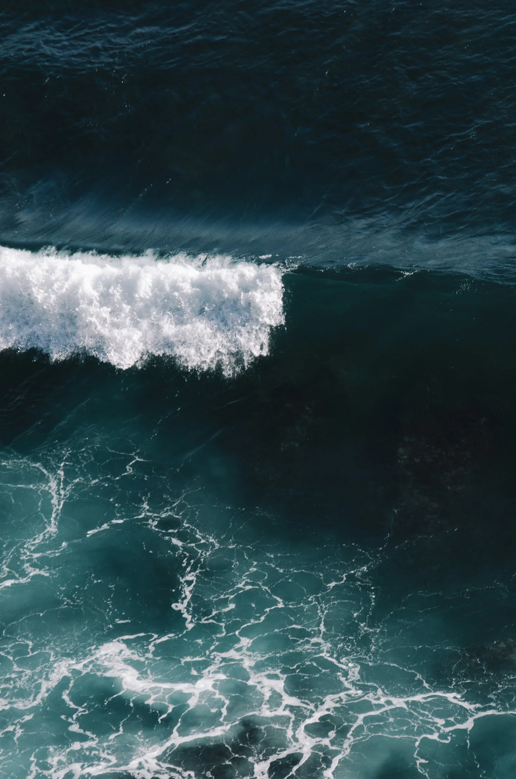 Aerial view of ocean waves with foam and ripples, showing deep blue and turquoise colors.