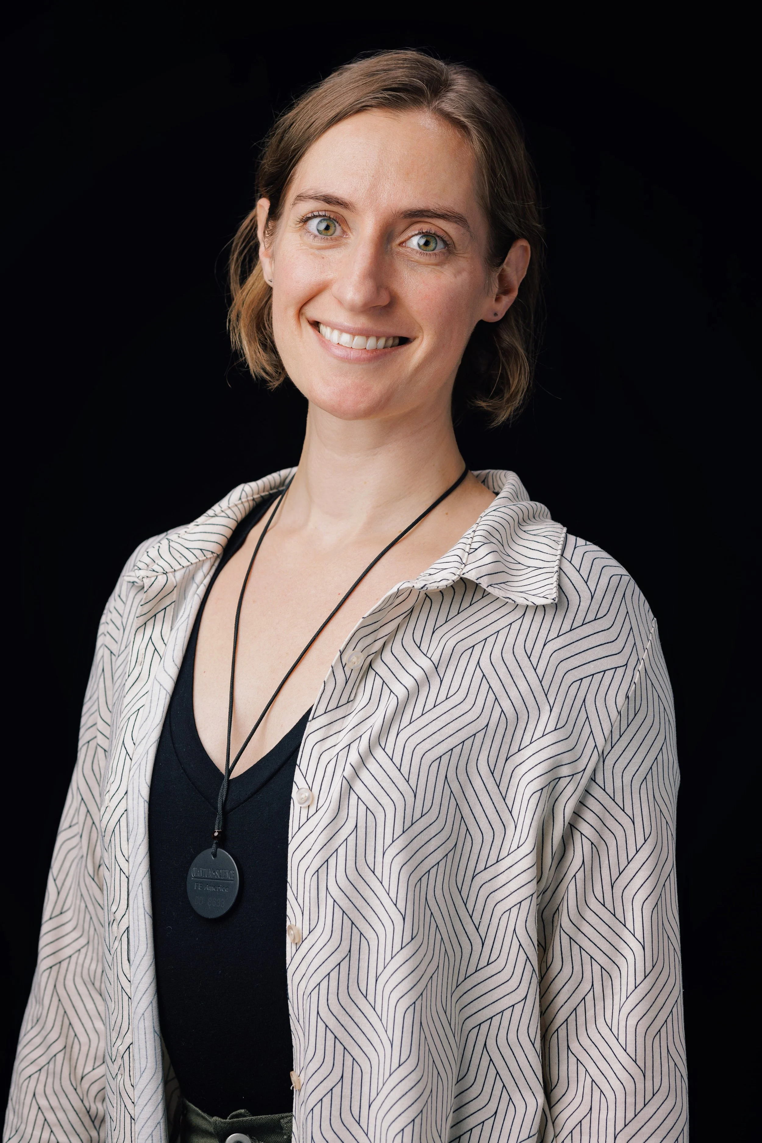 A woman with short brown hair and green eyes smiling, wearing a striped beige and white shirt over a black top, with a lanyard and badge, against a black background.