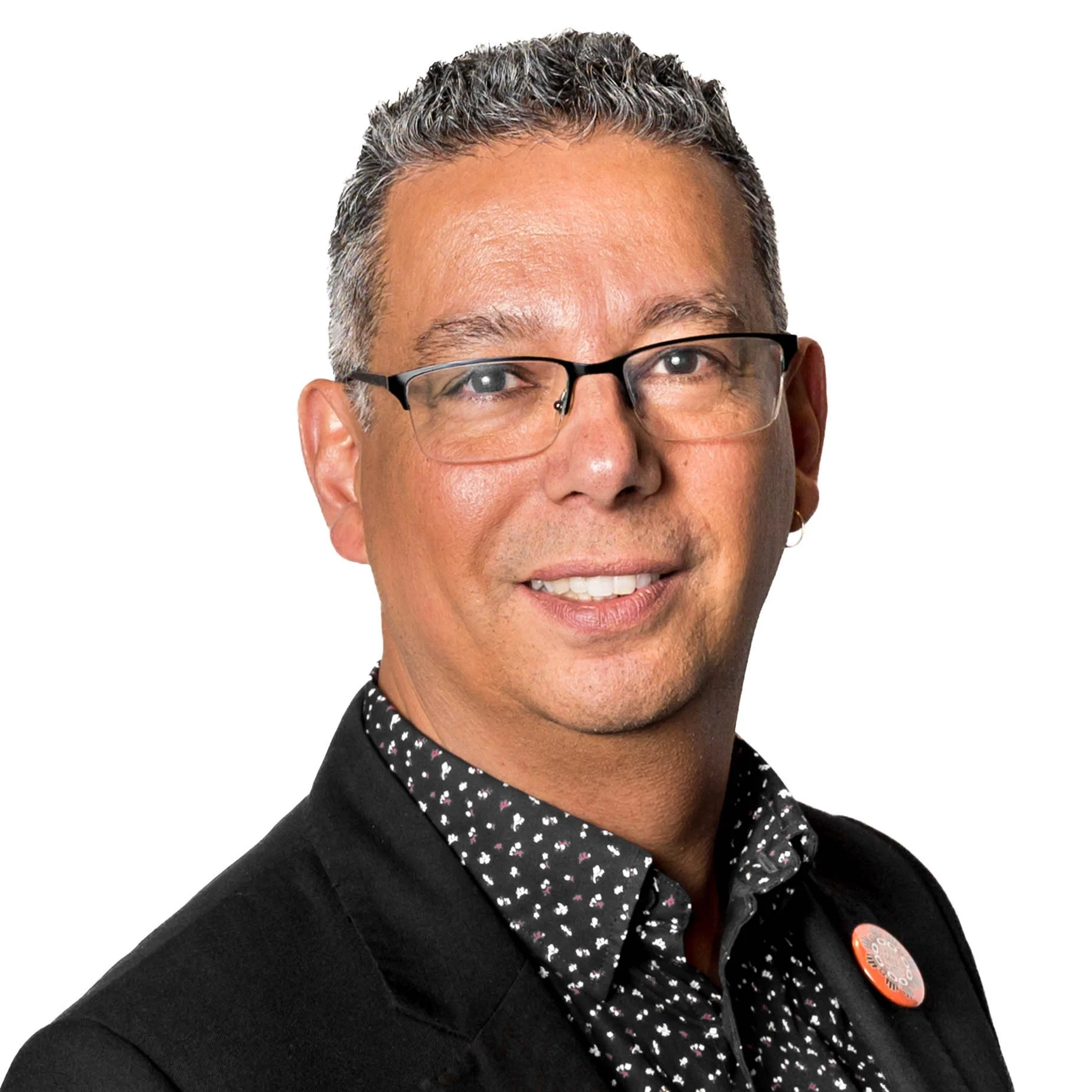 A close-up of Peter wearing black-rimmed glasses, a black blazer, and a black shirt with white and pink floral pattern, with short and curly gray hair, and a button on the blazer.