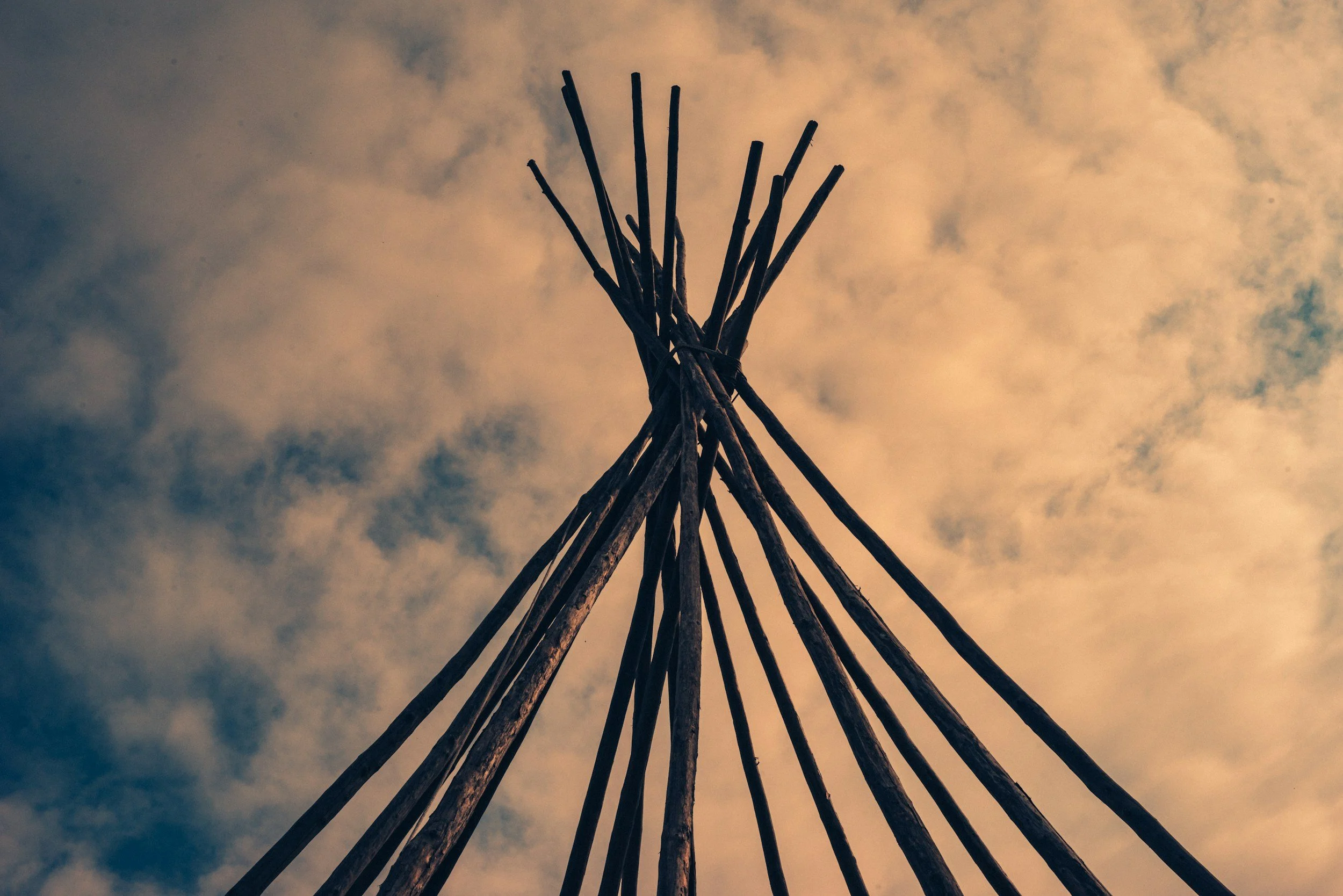 A structure made of wooden sticks arranged in a cone shape, viewed from below against a cloudy sky at sunset.