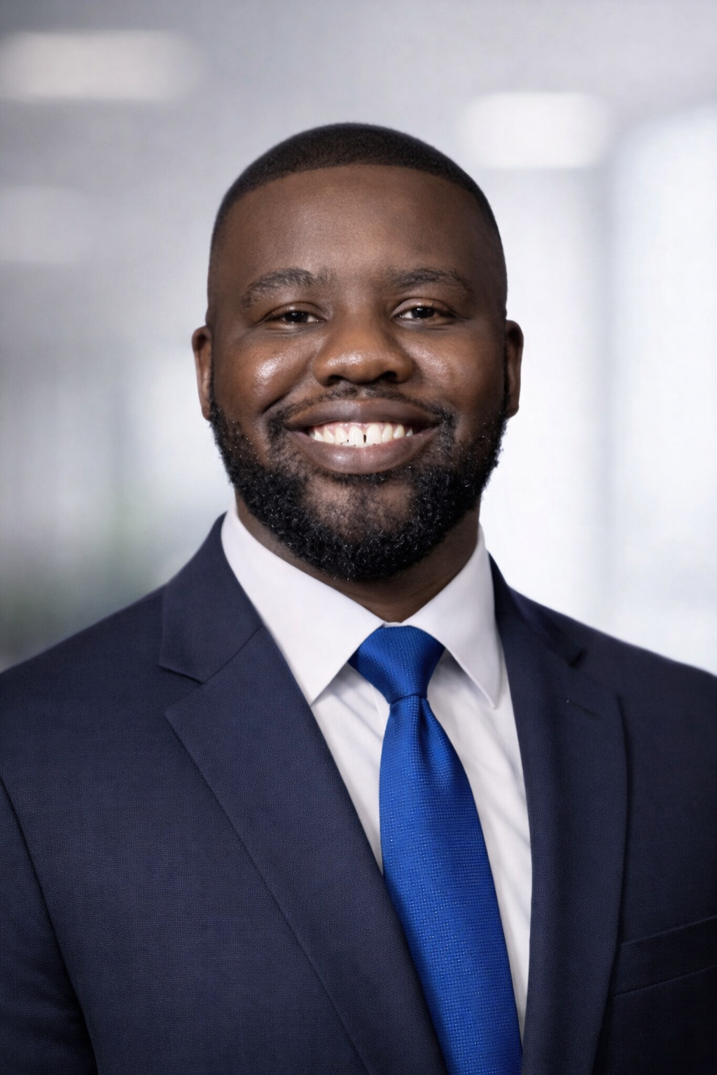 A smiling man with a beard wearing a dark suit, white shirt, and blue tie in an indoor setting.