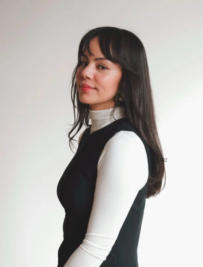 A woman with long dark hair wearing a white turtleneck and black sleeveless top, standing against a plain white background.