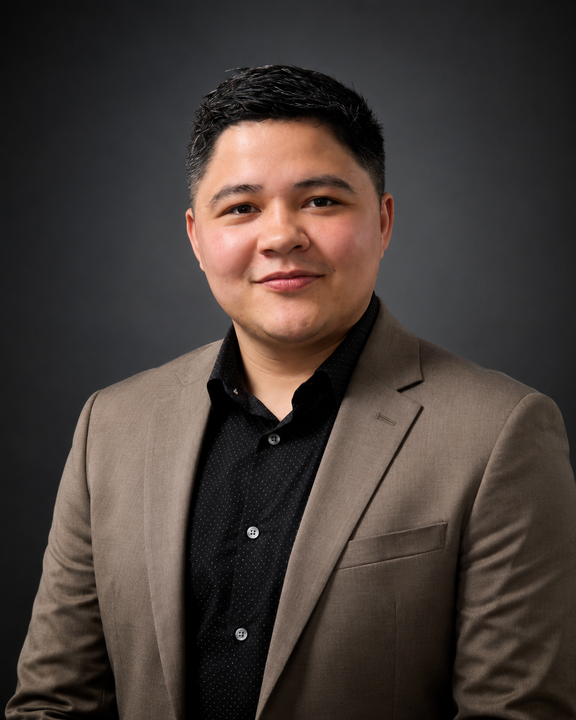 Business portrait of a young man with short black hair, wearing a brown suit jacket and black shirt, against a dark background.