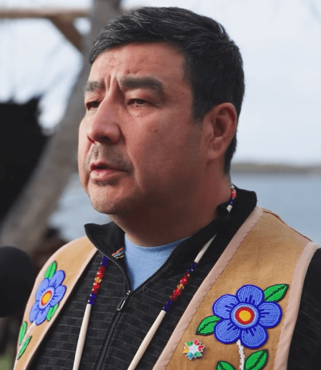 Close-up of a man wearing a tan vest with flower embroidery, speaking outdoors, near water, with a blurred background.