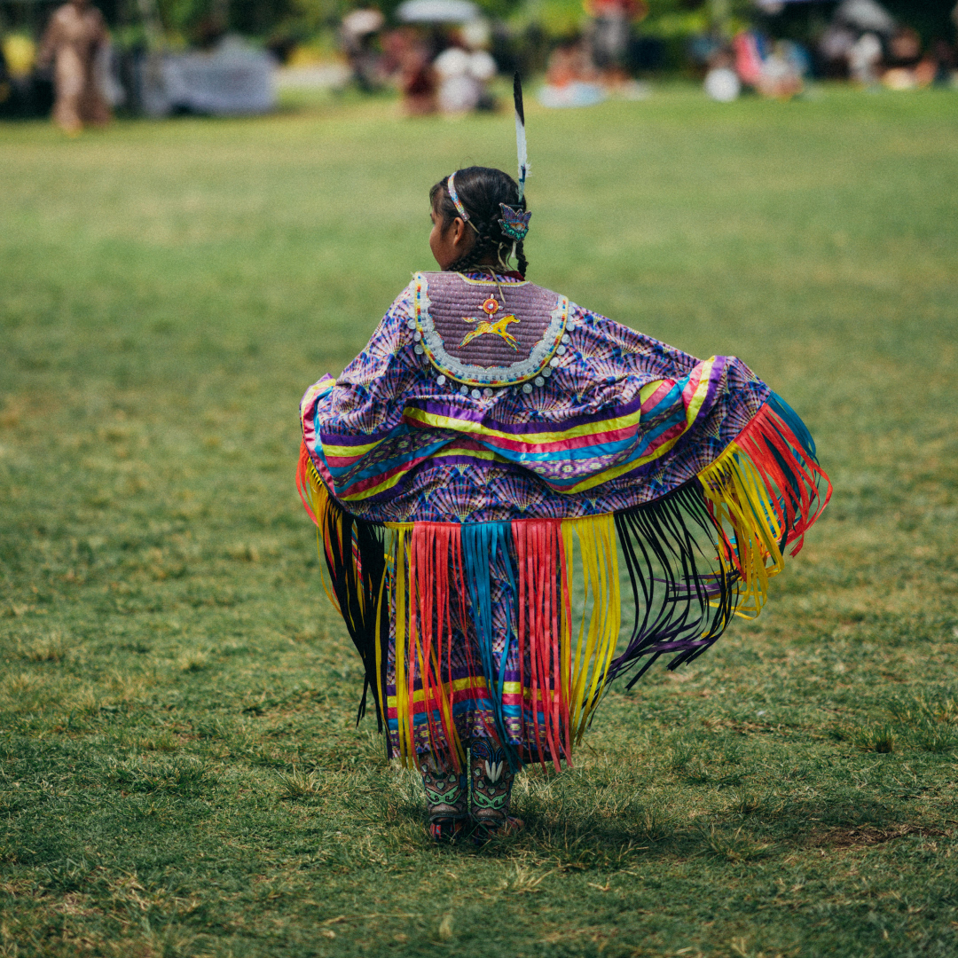 A young girl dressed in traditional, colorful Native American or indigenous attire, twirling in a grassy outdoor setting with blurred spectators in the background.