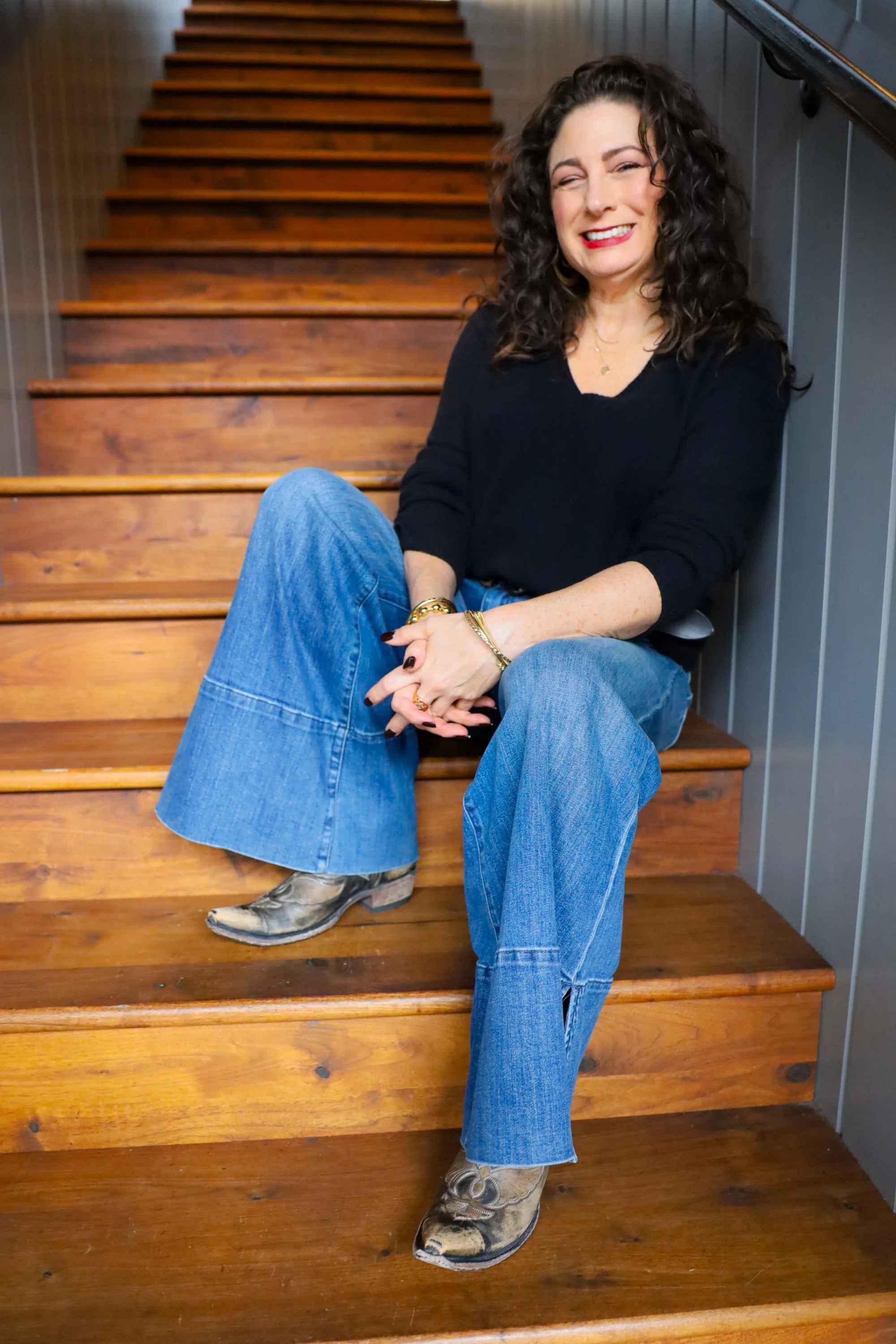 A woman therapist sitting on wooden stairs, wearing a black top, blue jeans, snakeskin cowboy boots, and gold jewelry. She is smiling and friendly-looking.