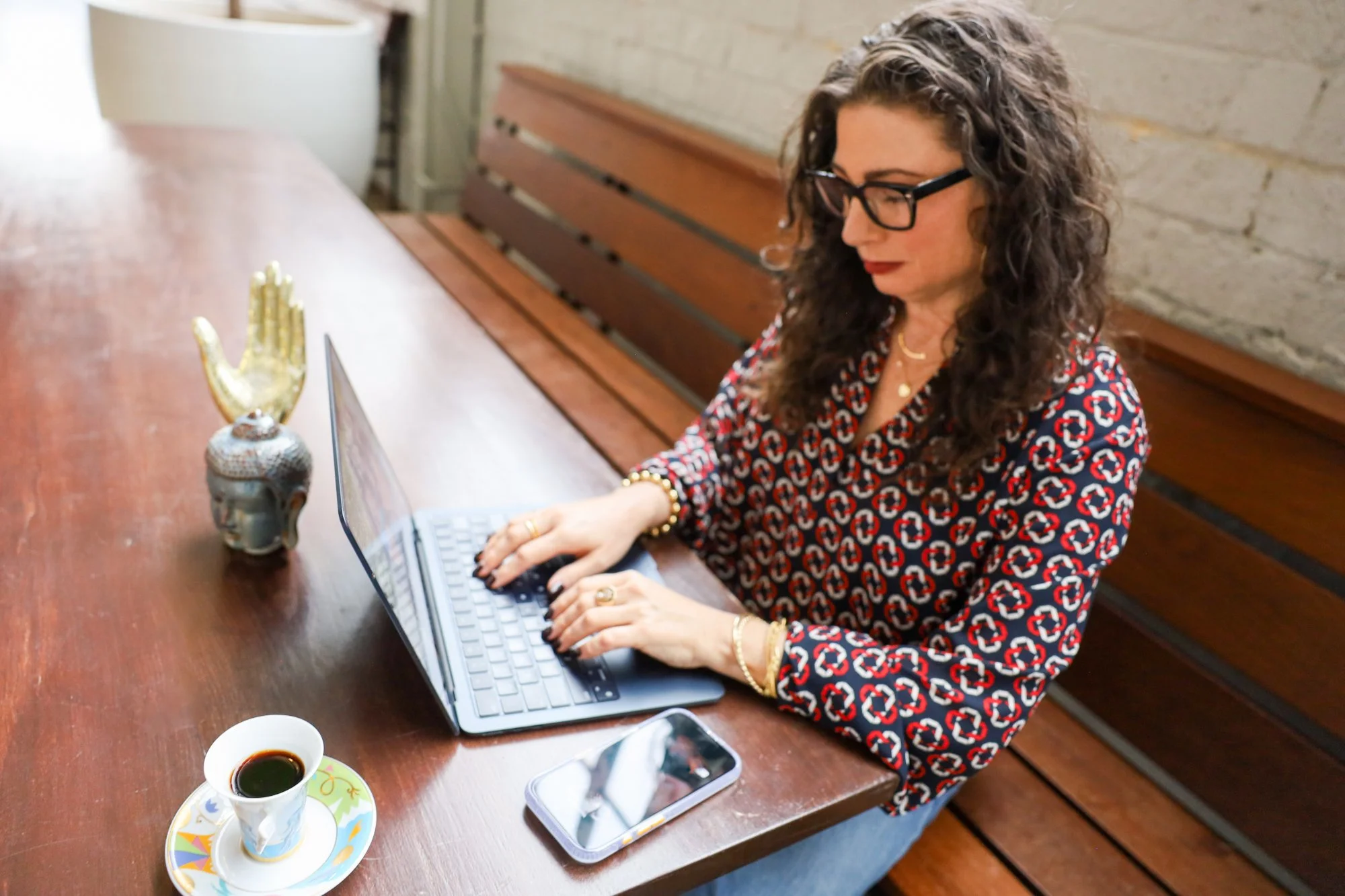 Woman therapist with curly hair and glasses working on a laptop at a wooden table, with a cup of coffee and decorative items on the table.