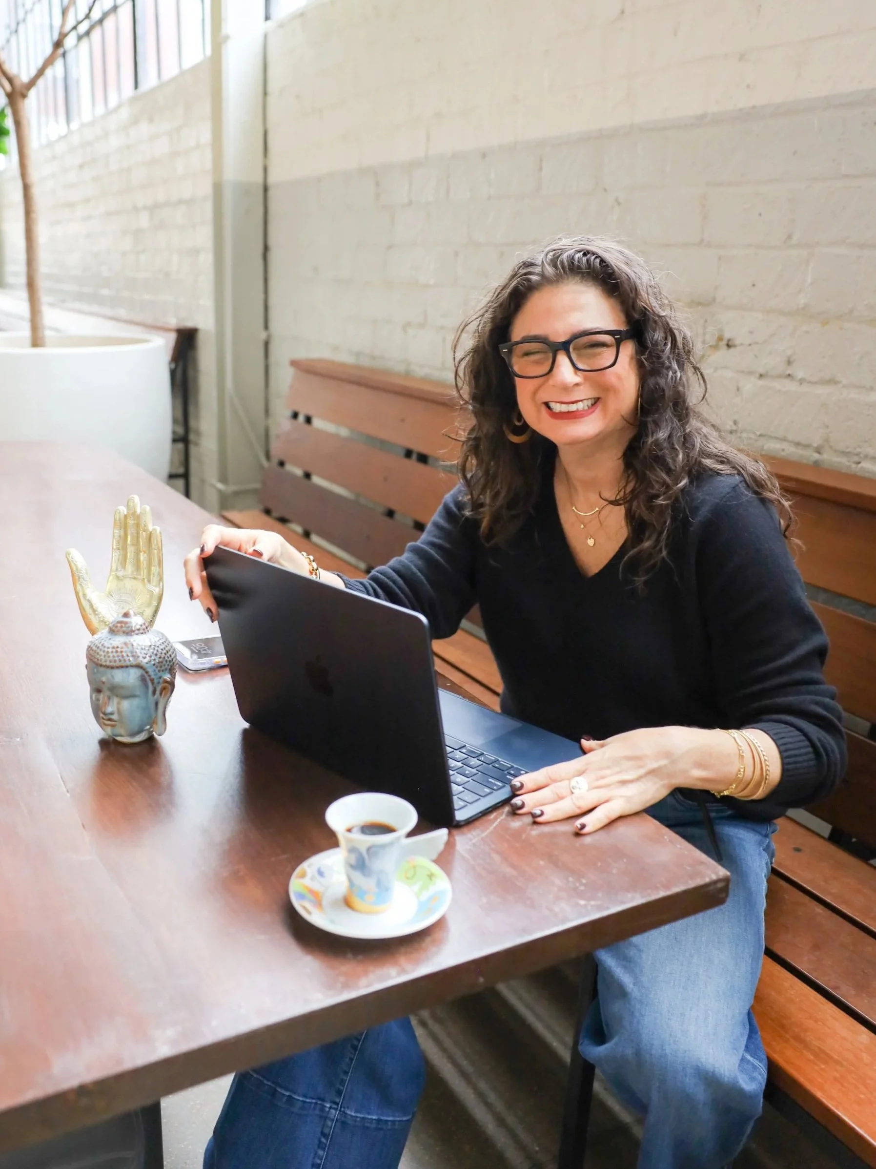 A woman therapist smiling while sitting at a wooden table with a laptop, coffee cup, and decorative items, in a well-lit cafe with a brick wall background.