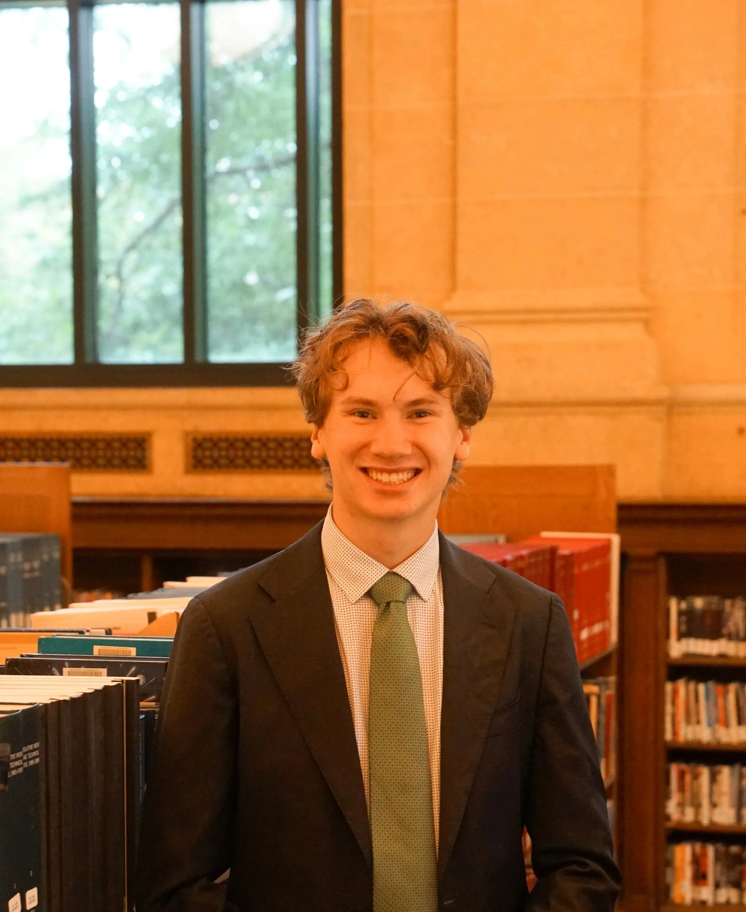 A young man in a dark suit, white shirt, and pale pink tie standing outside a building with classical columns and a staircase, smiling at the camera.