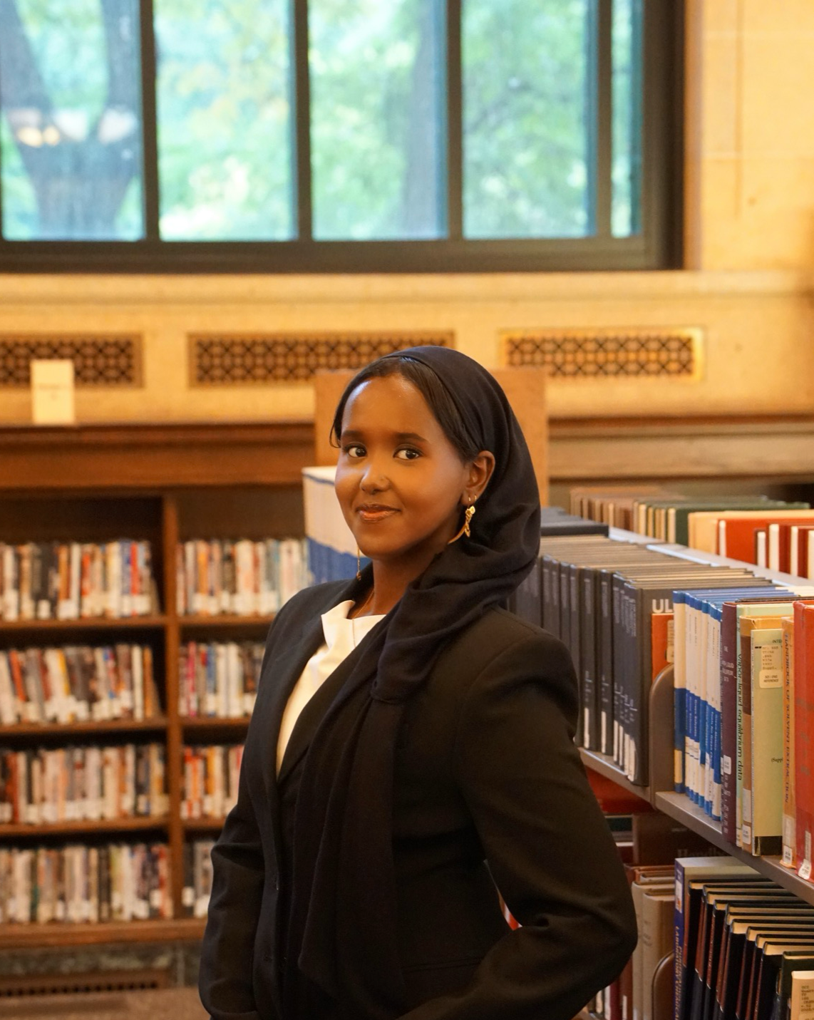 A young woman stands on the steps of a building with wooden doors in the background. She is wearing a black blazer, black skirt, and a black hijab, with her hands clasped in front of her.