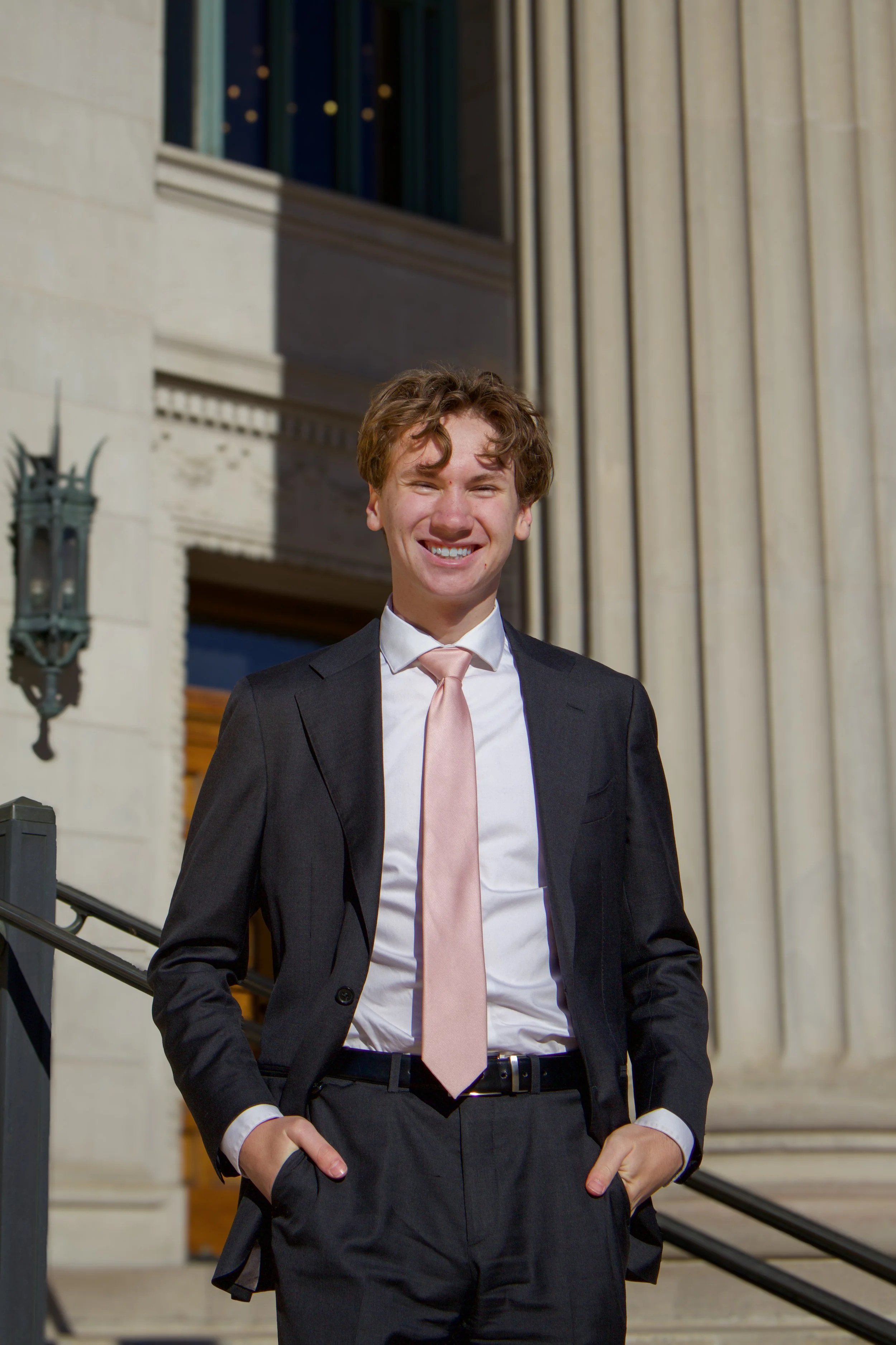 A young man in a dark suit, white shirt, and pale pink tie standing outside a building with classical columns and a staircase, smiling at the camera.