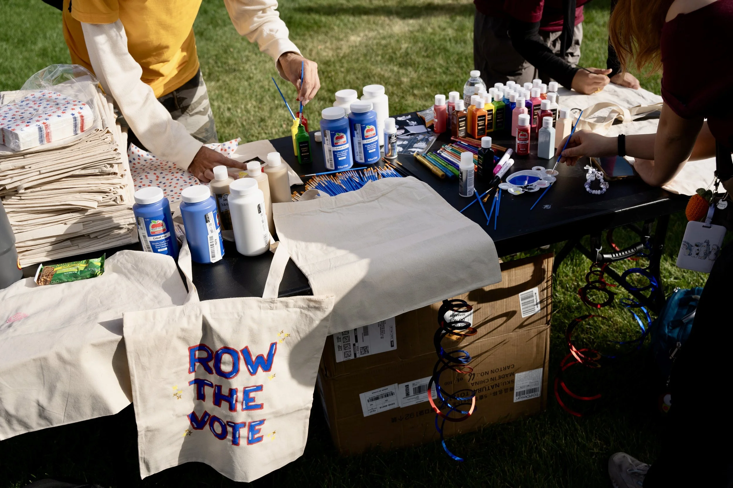 Table outdoors with supplies such as paint bottles, paintbrushes, paper bags, and a tote bag, possibly for voting or artistic activity, with people partially visible around the table.