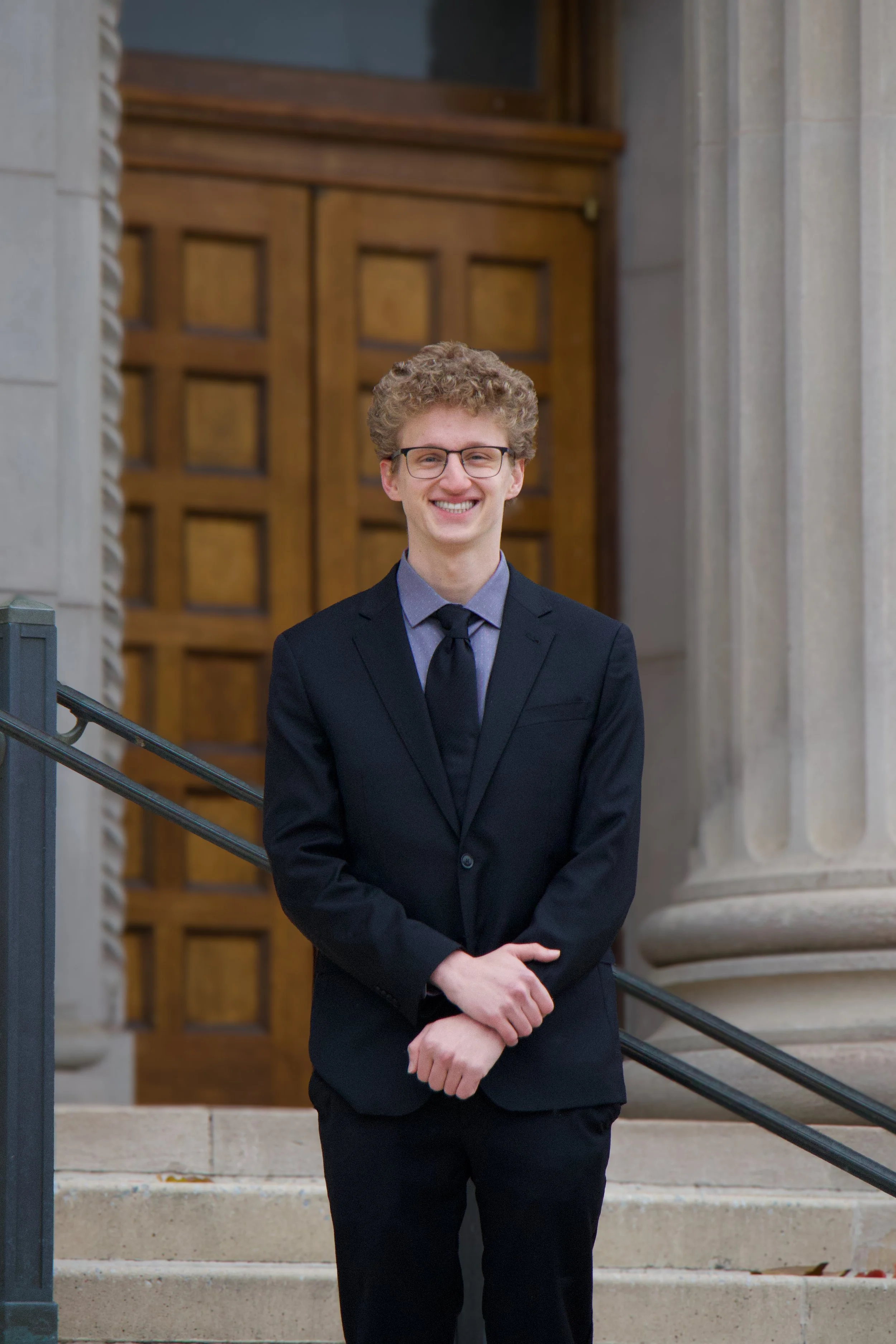 Young man with curly hair and glasses dressed in a dark suit, standing outside on steps in front of a stone building with a wooden door in the background.