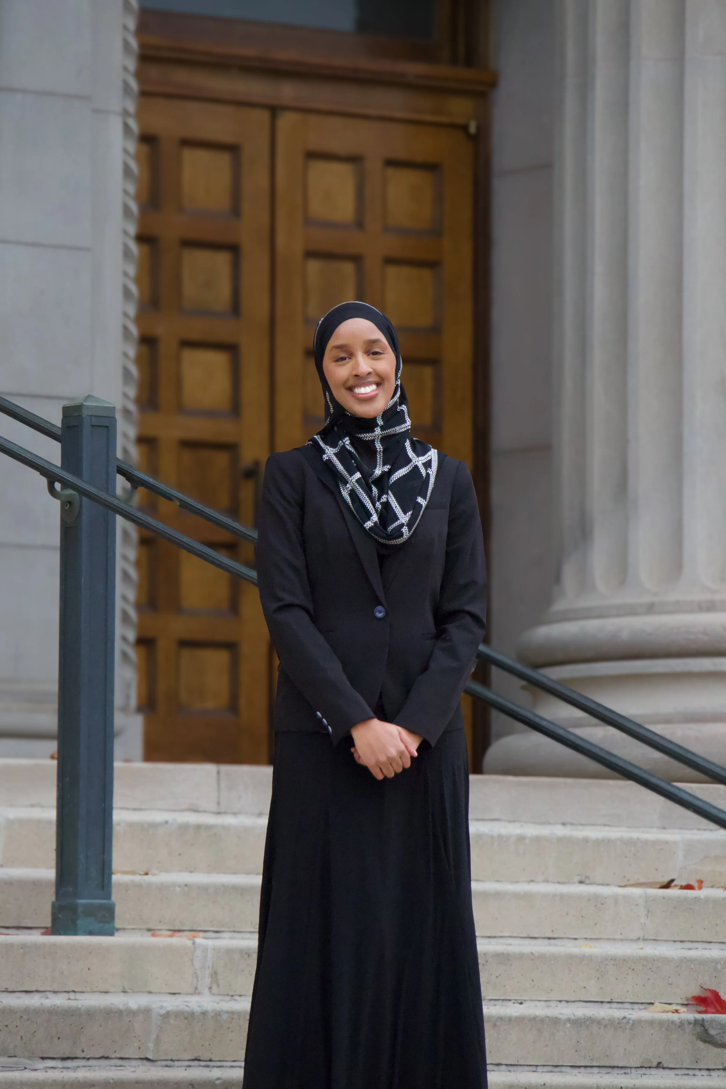 A woman wearing a black outfit and patterned headscarf standing on the steps of a building with wooden doors and stone columns.