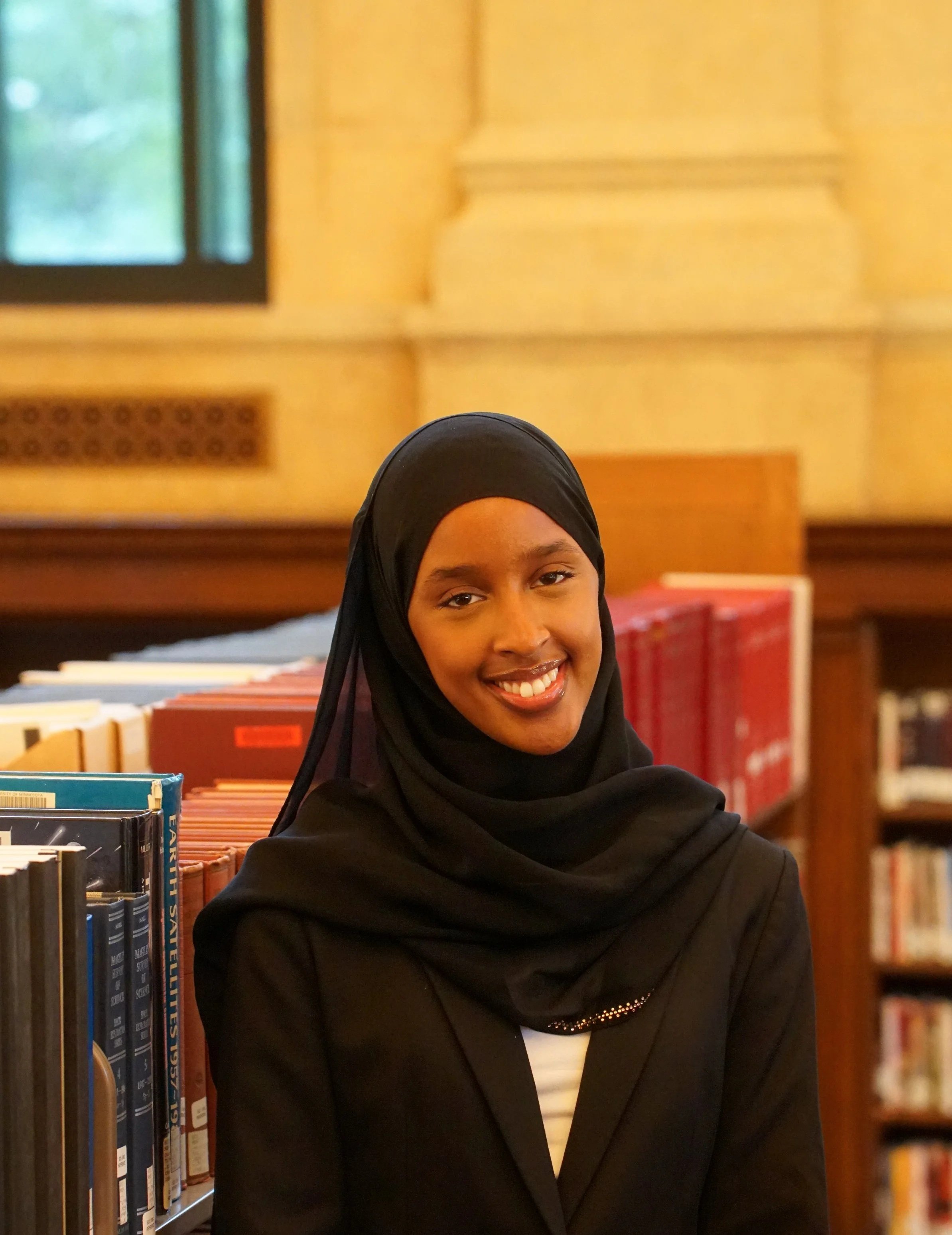 A woman wearing a black outfit and patterned headscarf standing on the steps of a building with wooden doors and stone columns.